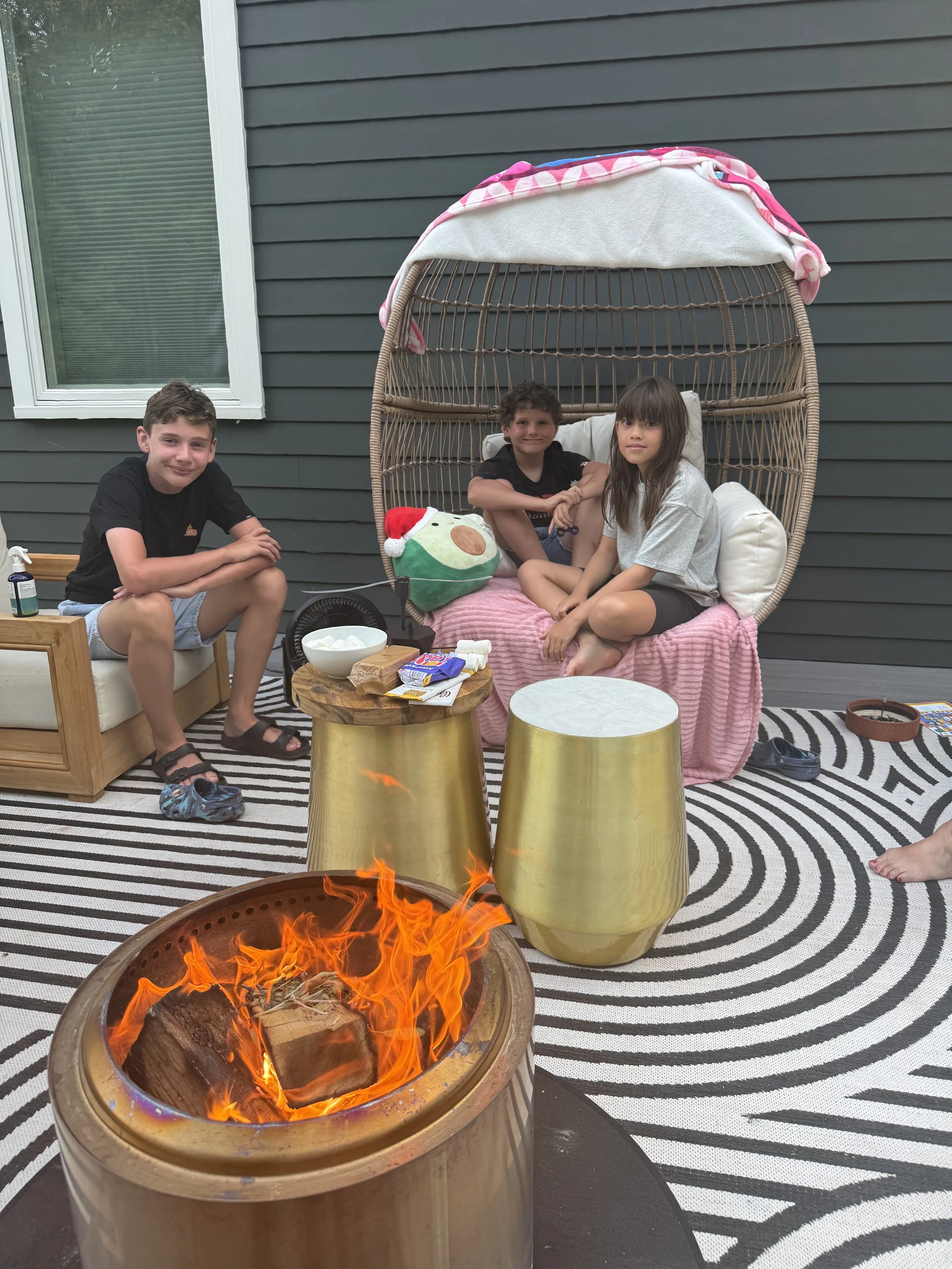 Three children sitting on a pink and white outdoor swing with a stuffed animal wearing a Santa hat, in a backyard with a fire pit, striped rug, small table with snacks, and a gray house wall in the background.