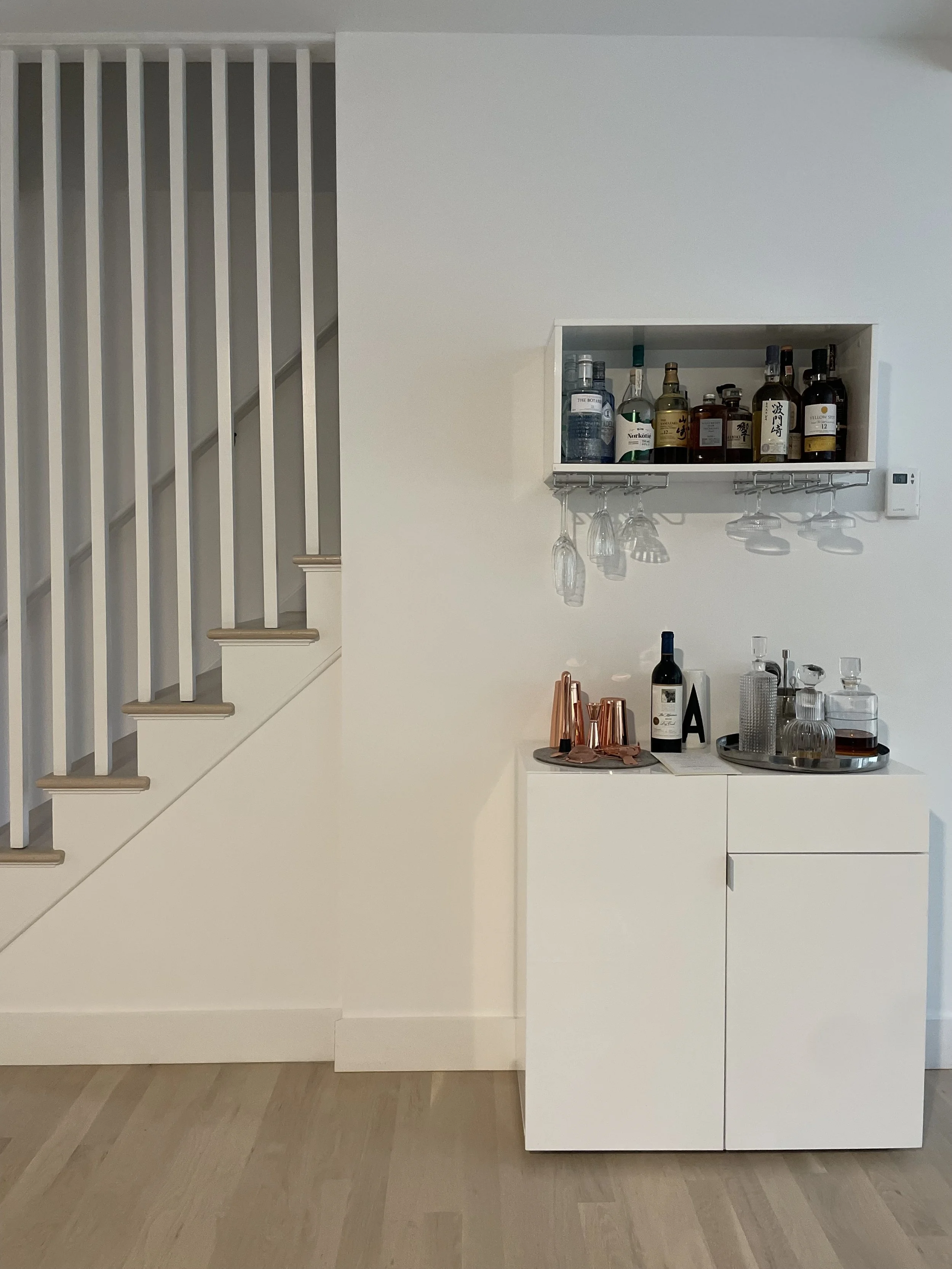 A white bar cabinet with various bottles of alcohol, glasses, and bar tools on top, positioned against a white wall near a staircase with white railings.