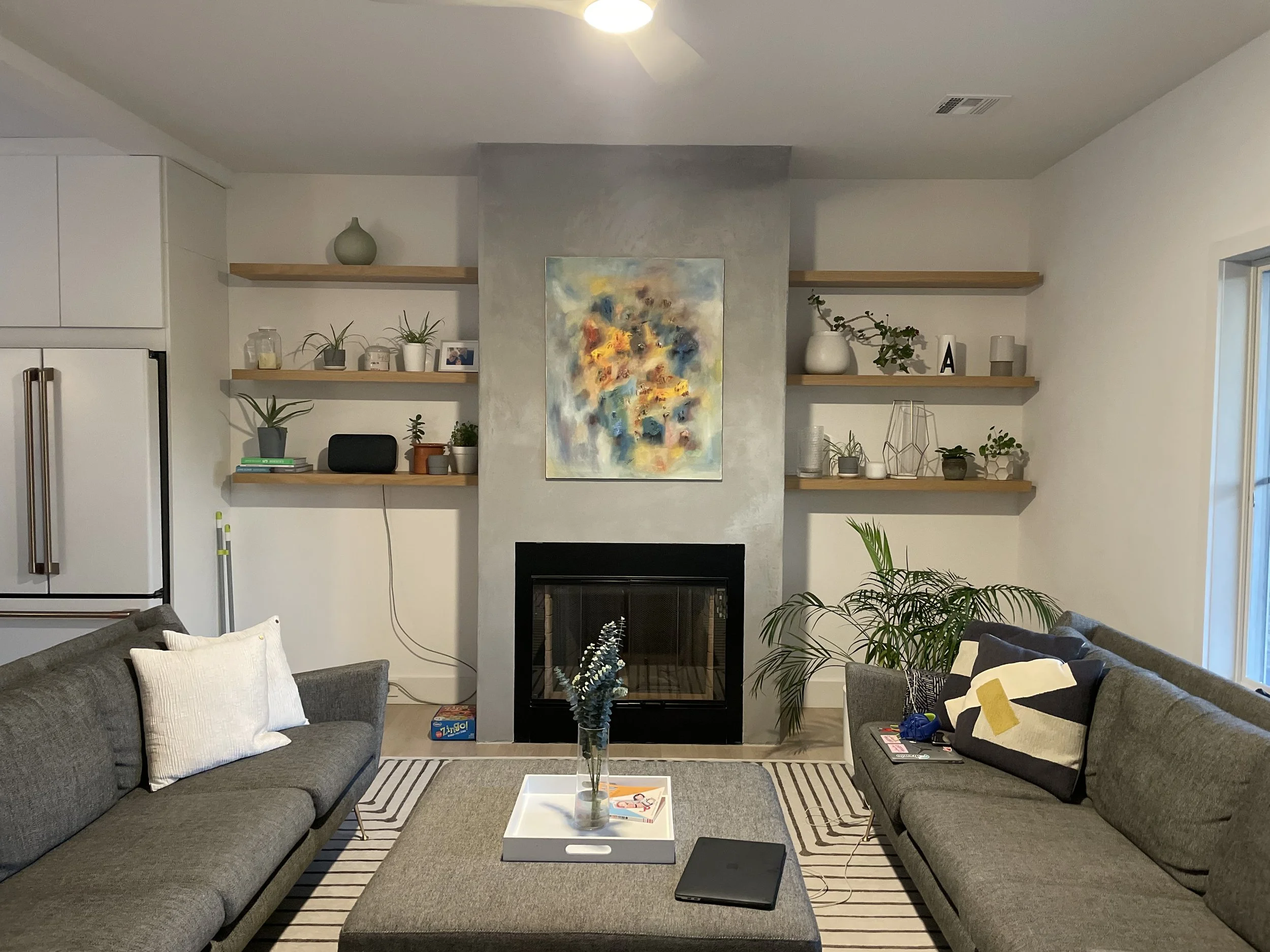 Living room with two gray sofas, a white tray on a large ottoman with a glass vase and books, a fireplace with colorful abstract art above, open wooden shelves with decorative items, houseplants, and a window on the right.