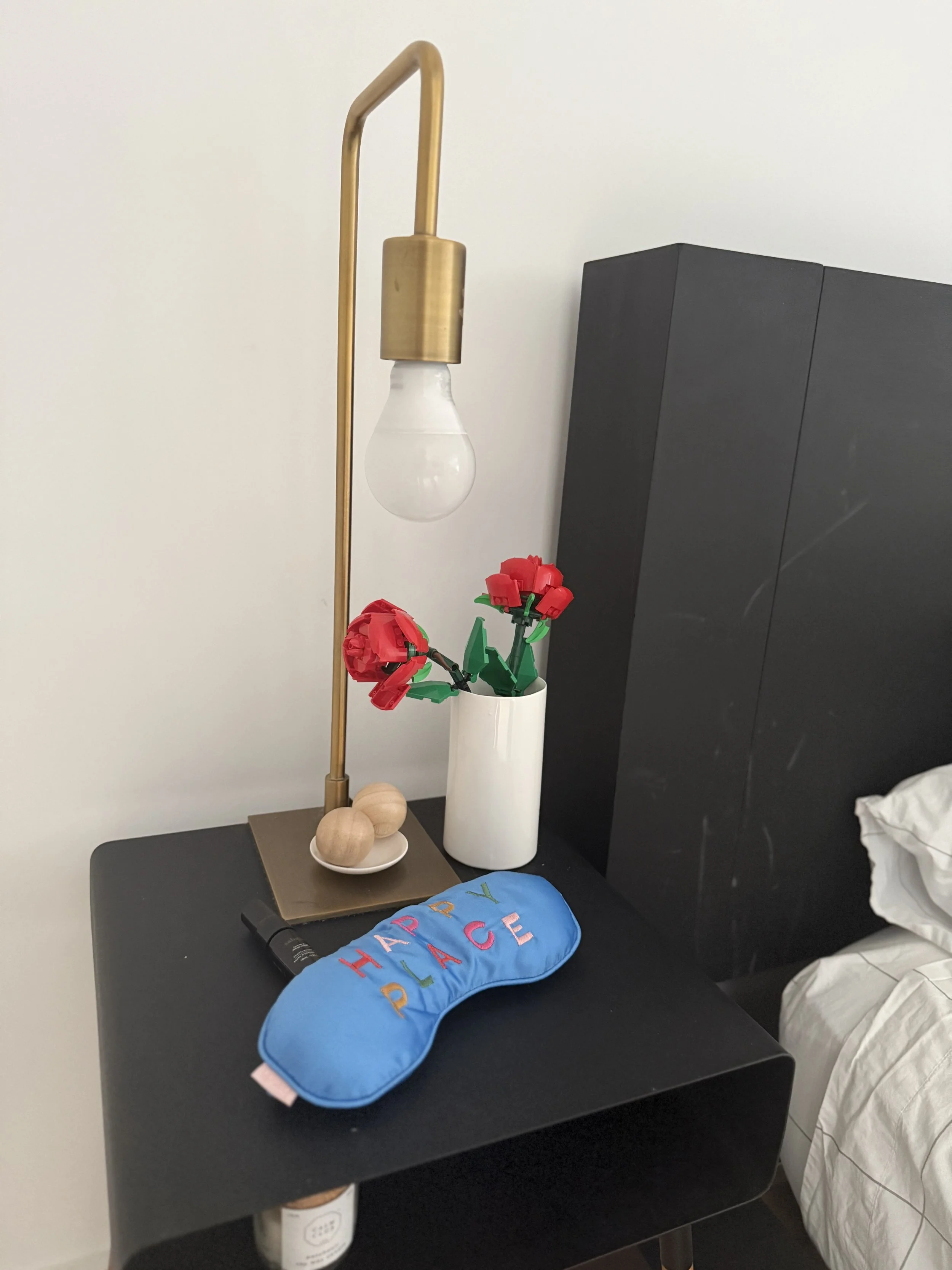 Nightstand with decorative items including a blue sleep mask reading "HAPPY PLACE," a black flashlight, a small dish with two wooden balls, a white vase with red flowers, a brass-colored lamp without a light bulb, and a black headboard next to a whit