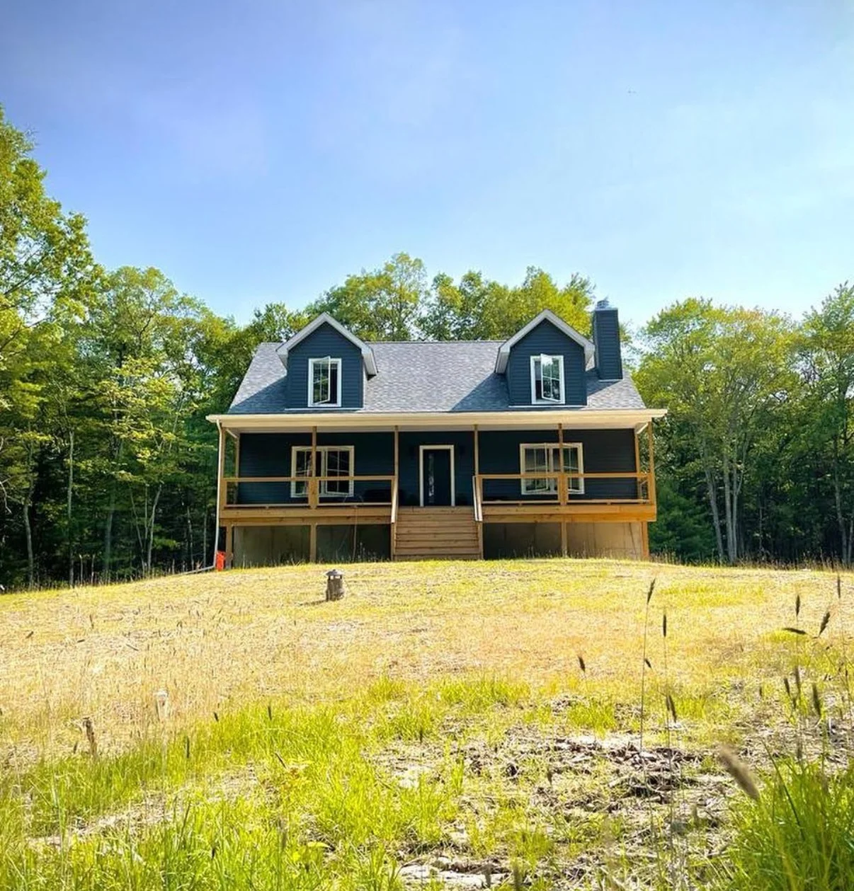 A two-story house with a dark blue exterior, large front porch, and two dormer windows on the roof, situated in a clearing surrounded by green trees and a grassy lawn.