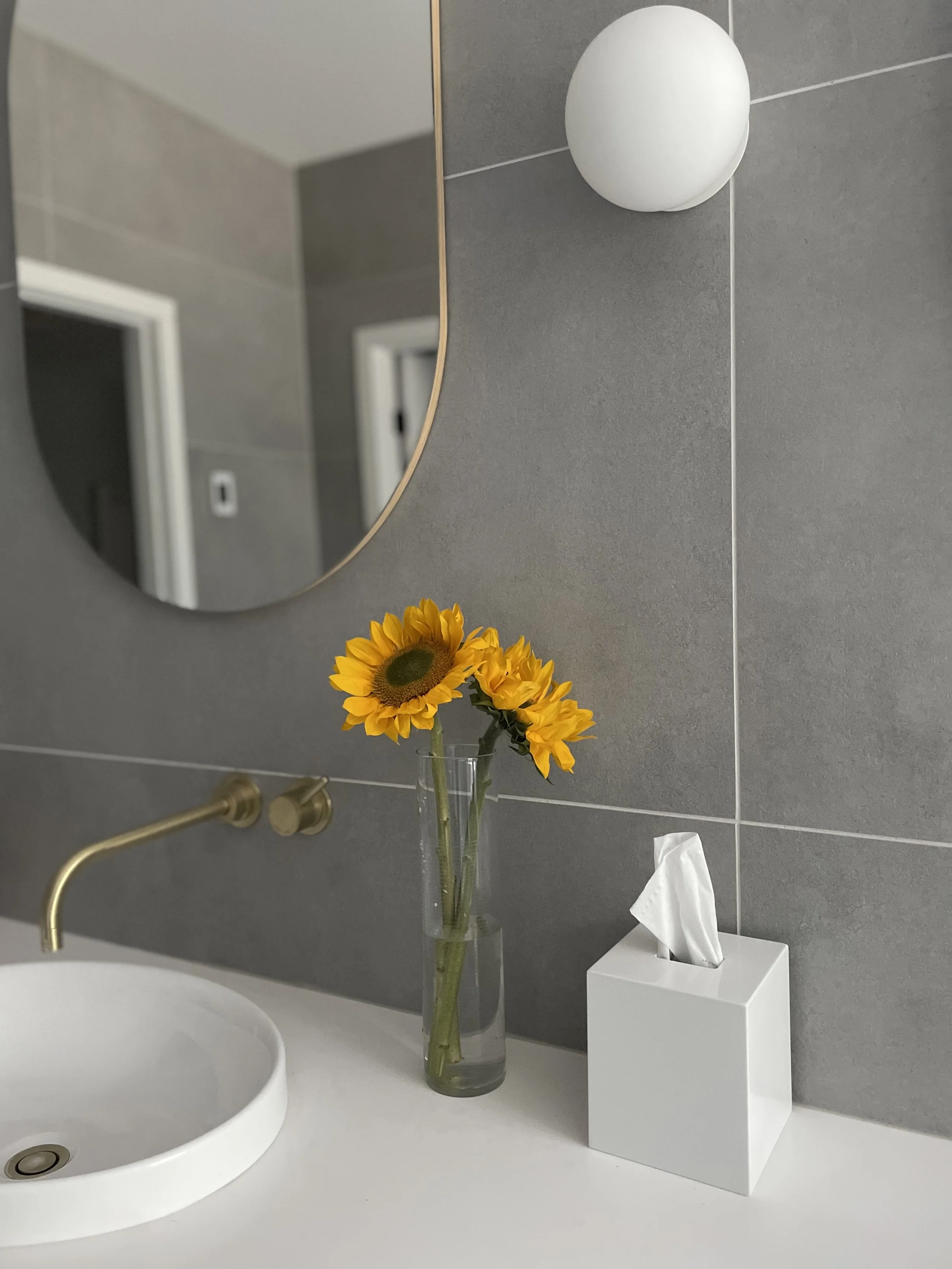 A bathroom vanity with a white sink, brass faucet, a clear glass vase with sunflowers, and a white tissue box, with a wall mirror and gray tiled wall.