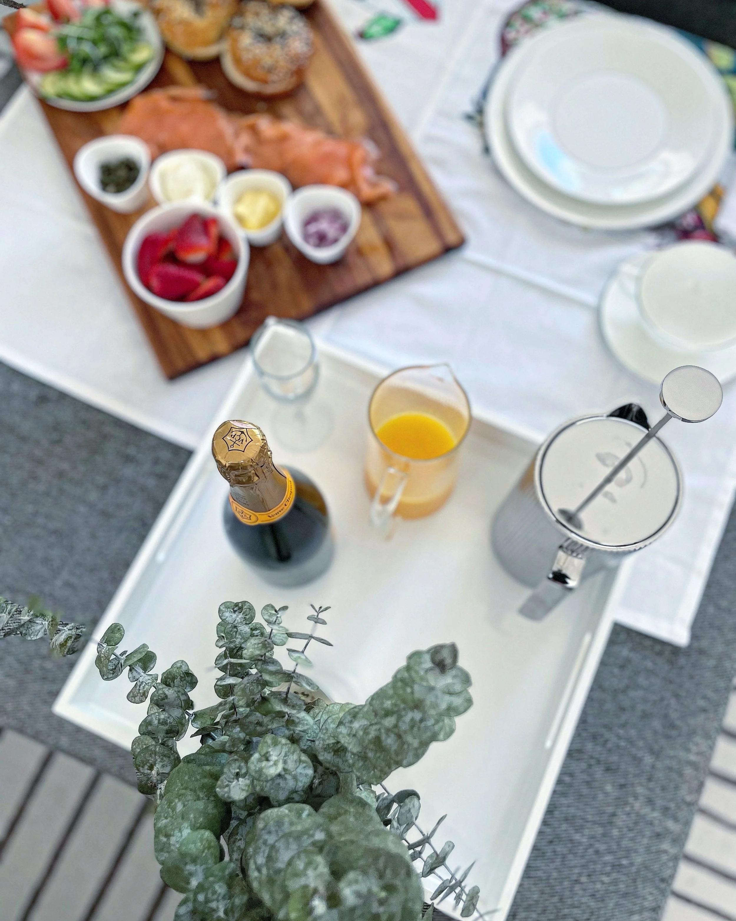 Table set for a brunch, featuring a tray with muffins, fruit, and condiments, a pitcher of orange juice, a bottle of champagne, a glass with orange juice, and stacked white plates, with a green plant in the foreground.