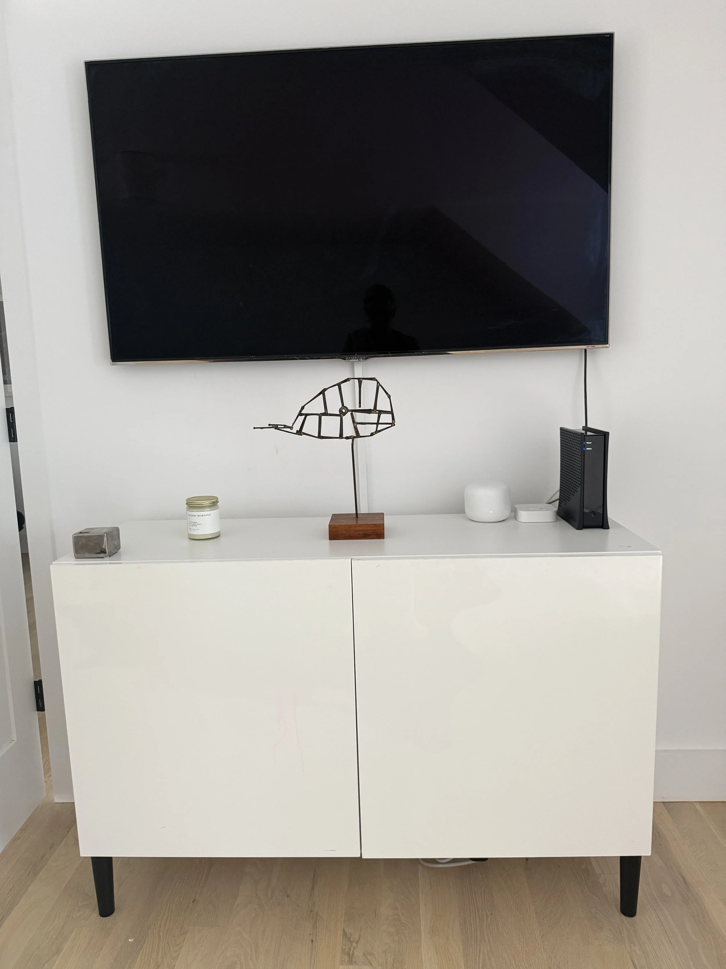 White cabinet with black legs and a flat top, holding decorative items including a wire and wood sculpture, a candle, a small stone, a white rounded container, and a black electronic device, against a white wall with a large black TV mounted above.