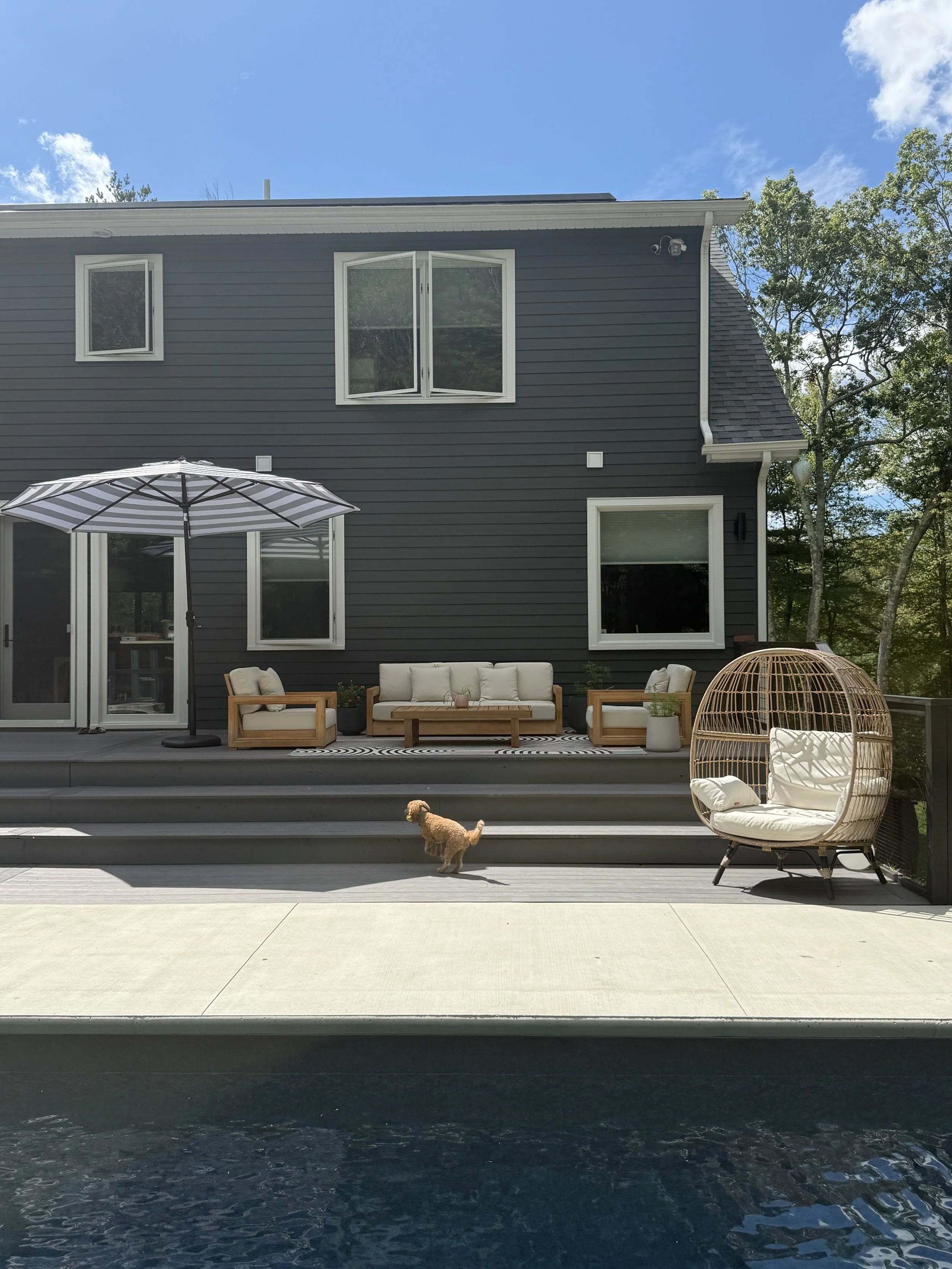 Backyard patio with outdoor furniture, including a sofa, two chairs, and a large parasol, against a dark gray house with large windows, surrounded by trees and a small dog walking on the patio.