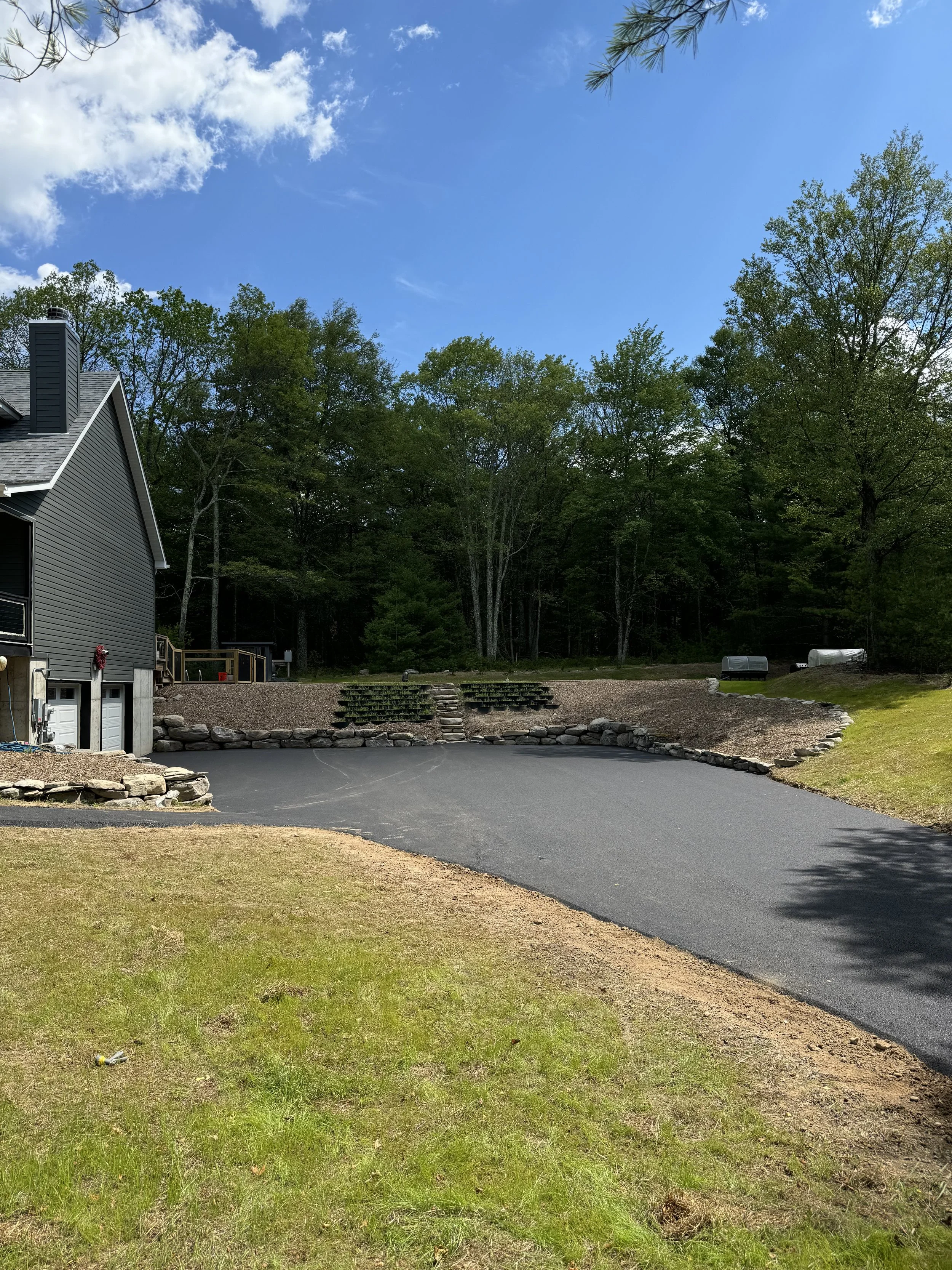 Freshly paved black asphalt driveway in backyard with a grassy lawn on the sides, a house on the left, and a wooded area in the background under a blue sky with some clouds.