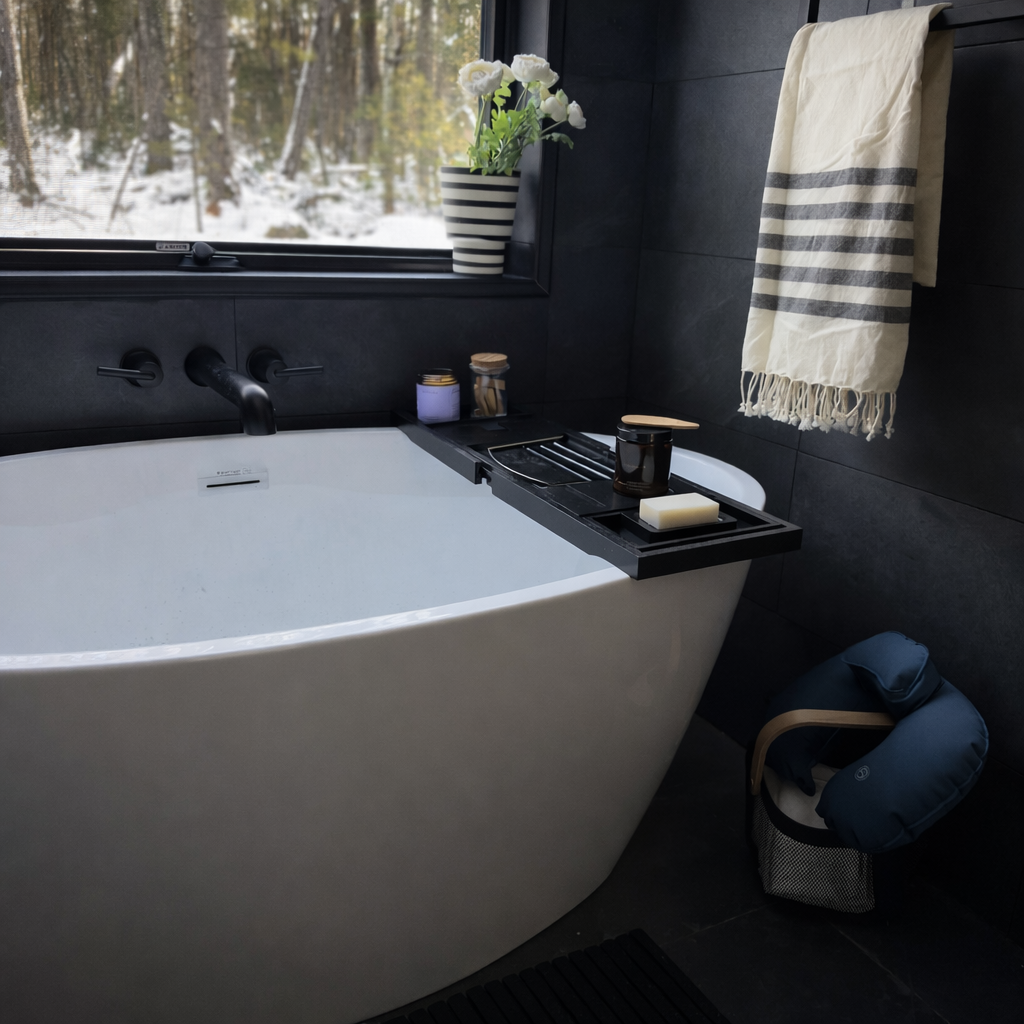 Bathroom with a white bathtub, black wall tiles, a window with a snowy forest view, a black shelf holding a bar of soap, a candle, and various luxury items for a relaxing bath.