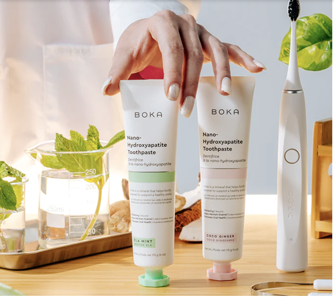 Person holding two tubes of BOKA Nano-Hydroxyapatite Toothpaste in a bright, clean setting with dental tools and green leaves nearby.