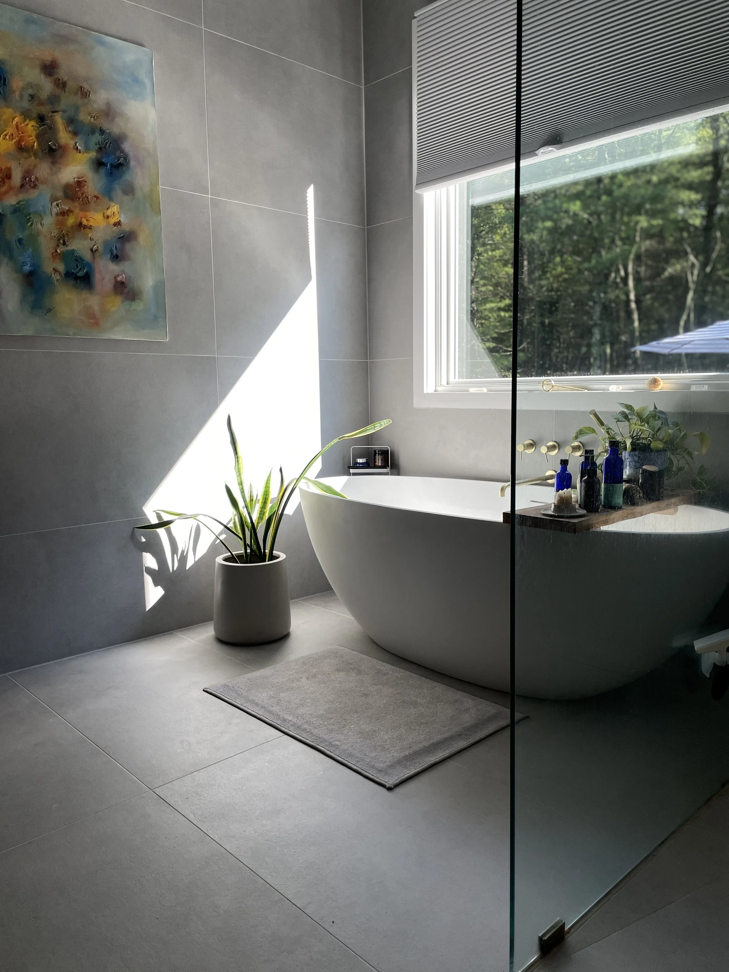 Modern bathroom with a white freestanding bathtub, a potted plant, and an abstract painting on the wall. Natural light streams through a large window, creating a shadow on the wall.