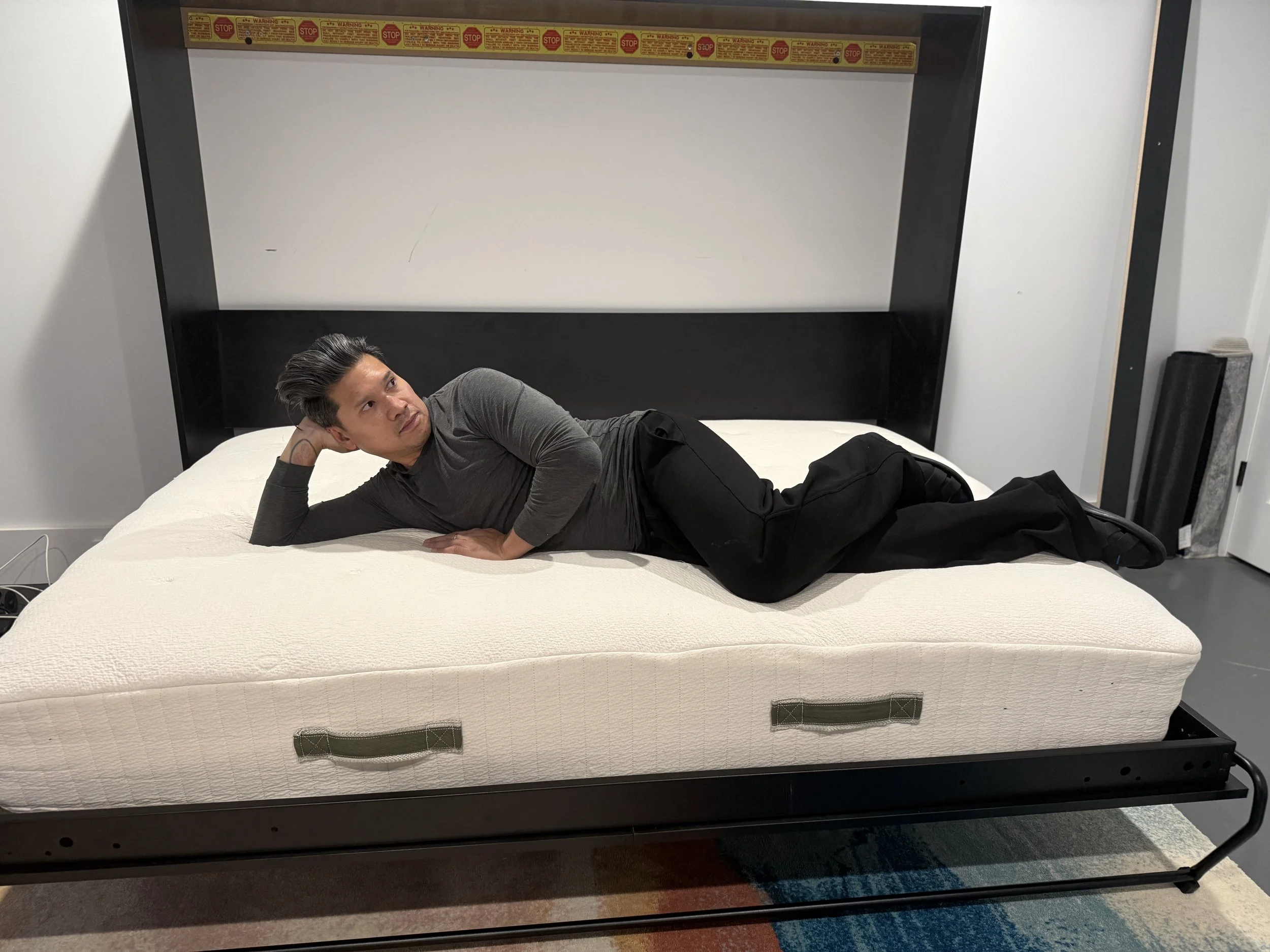 A man laying on a mattress, propped up on one elbow, in a room with plain walls and a carpeted floor.
