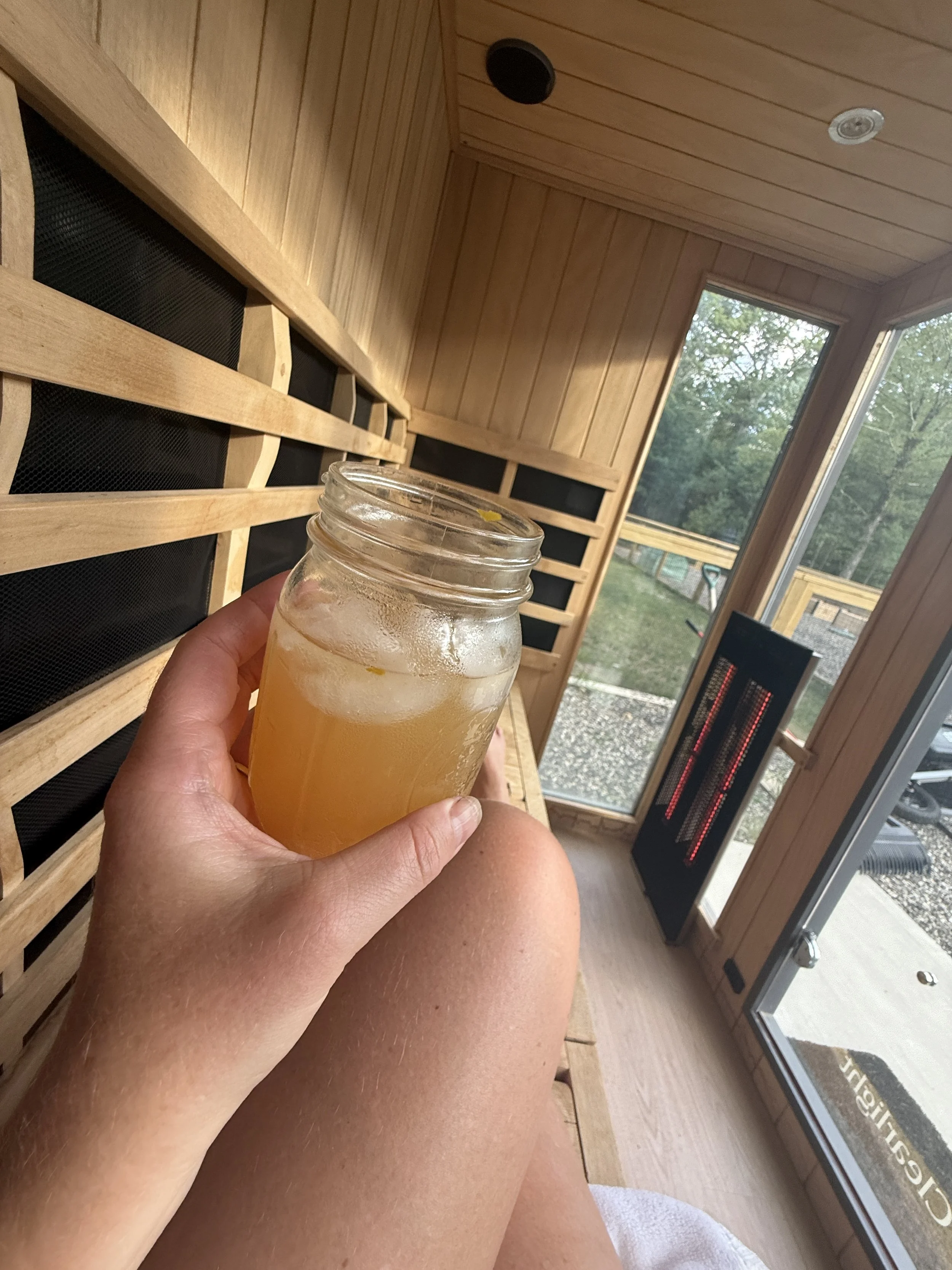 Person holding a mason jar filled with a cold, frothy beverage inside a wooden sauna with large windows showing greenery outside.