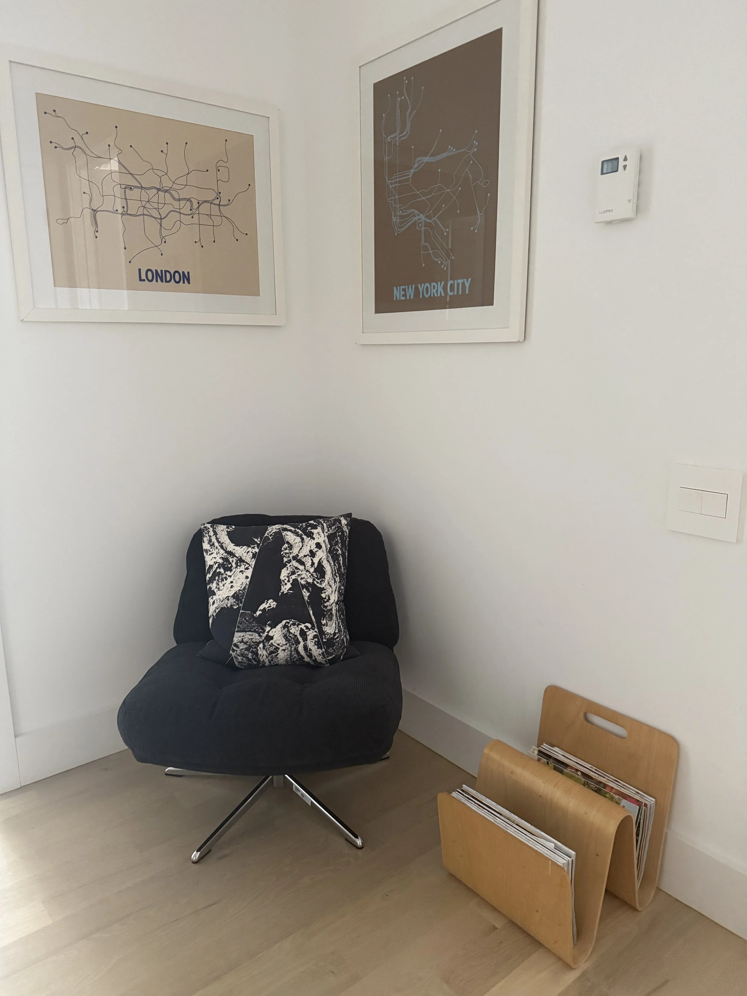 A cozy corner with a black swivel chair that has a black and white patterned pillow on it, next to a wooden magazine rack filled with magazines, and two framed maps of London and New York City on the white wall.