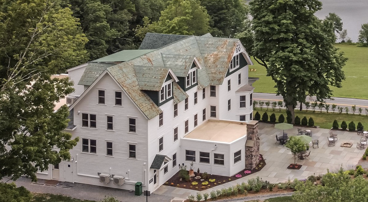 A large white multi-story house with a porch and a rusted metal roof, surrounded by green trees and a landscaped outdoor patio area with tables, chairs, and umbrellas.
