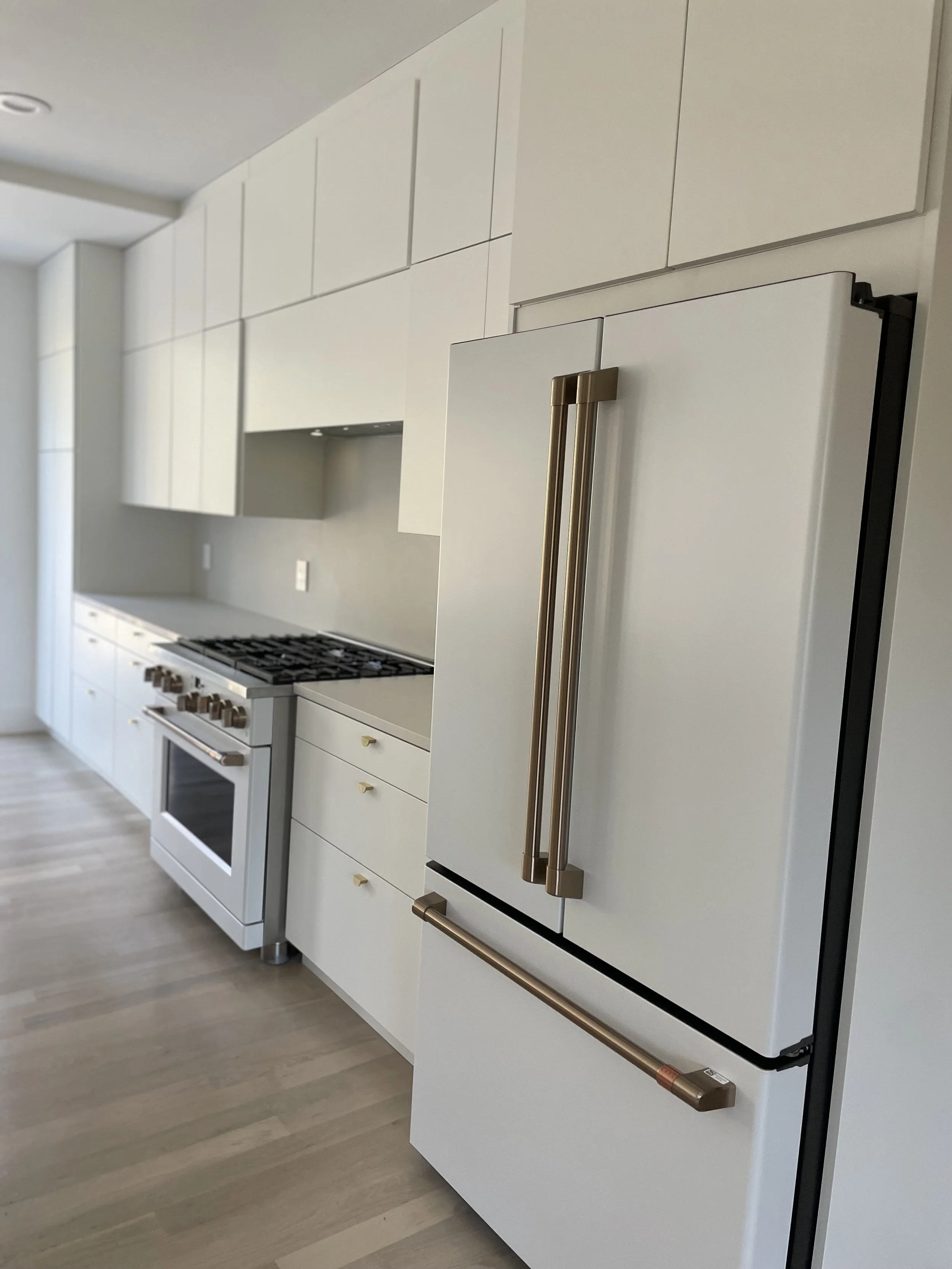 Modern white kitchen with gold handles on cabinets and appliances, including a refrigerator, stove, and oven, with light wood flooring.