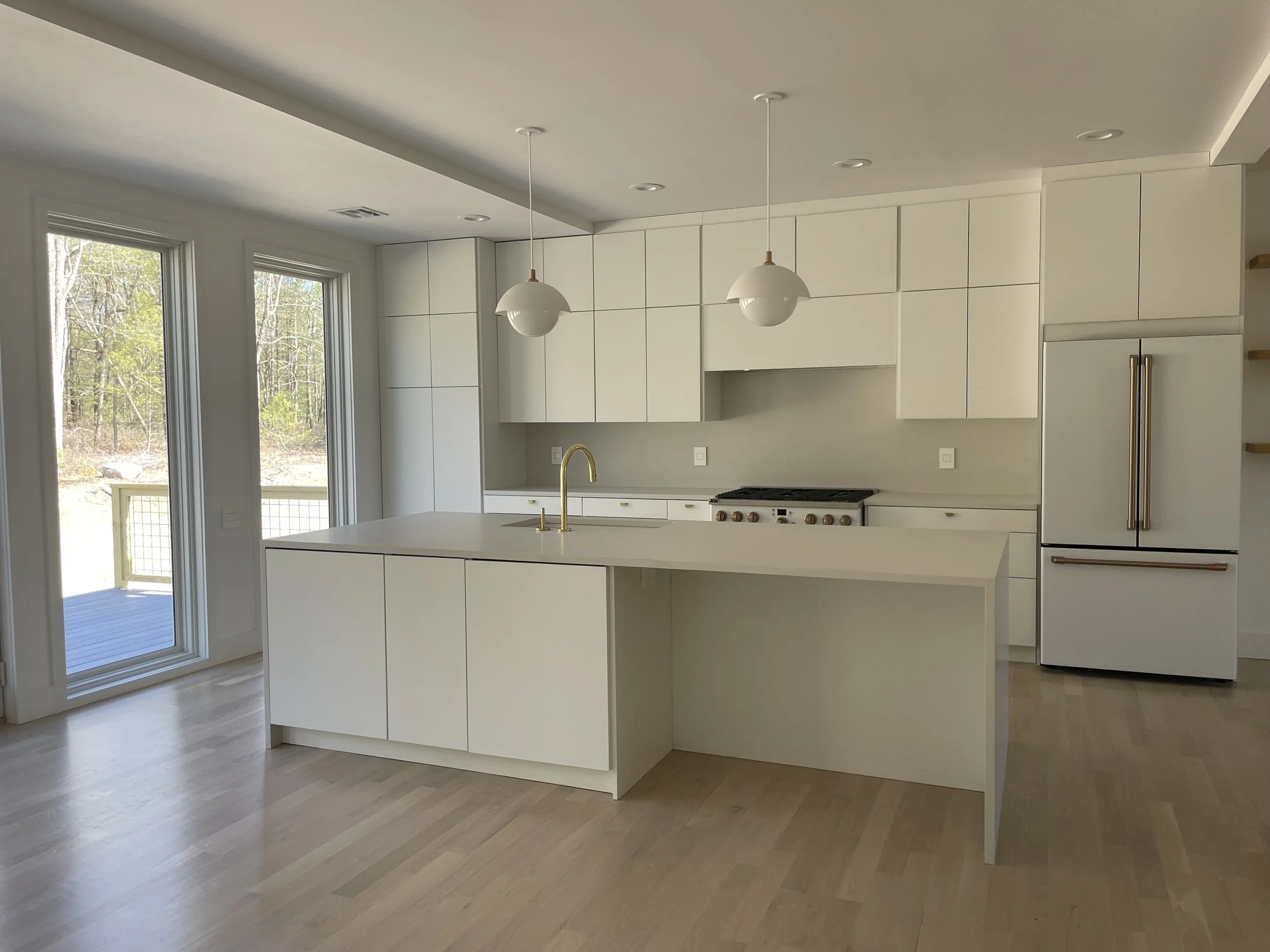 Modern white kitchen with large island, double glass doors leading outside, white cabinetry, gold fixtures, and a stainless steel refrigerator.