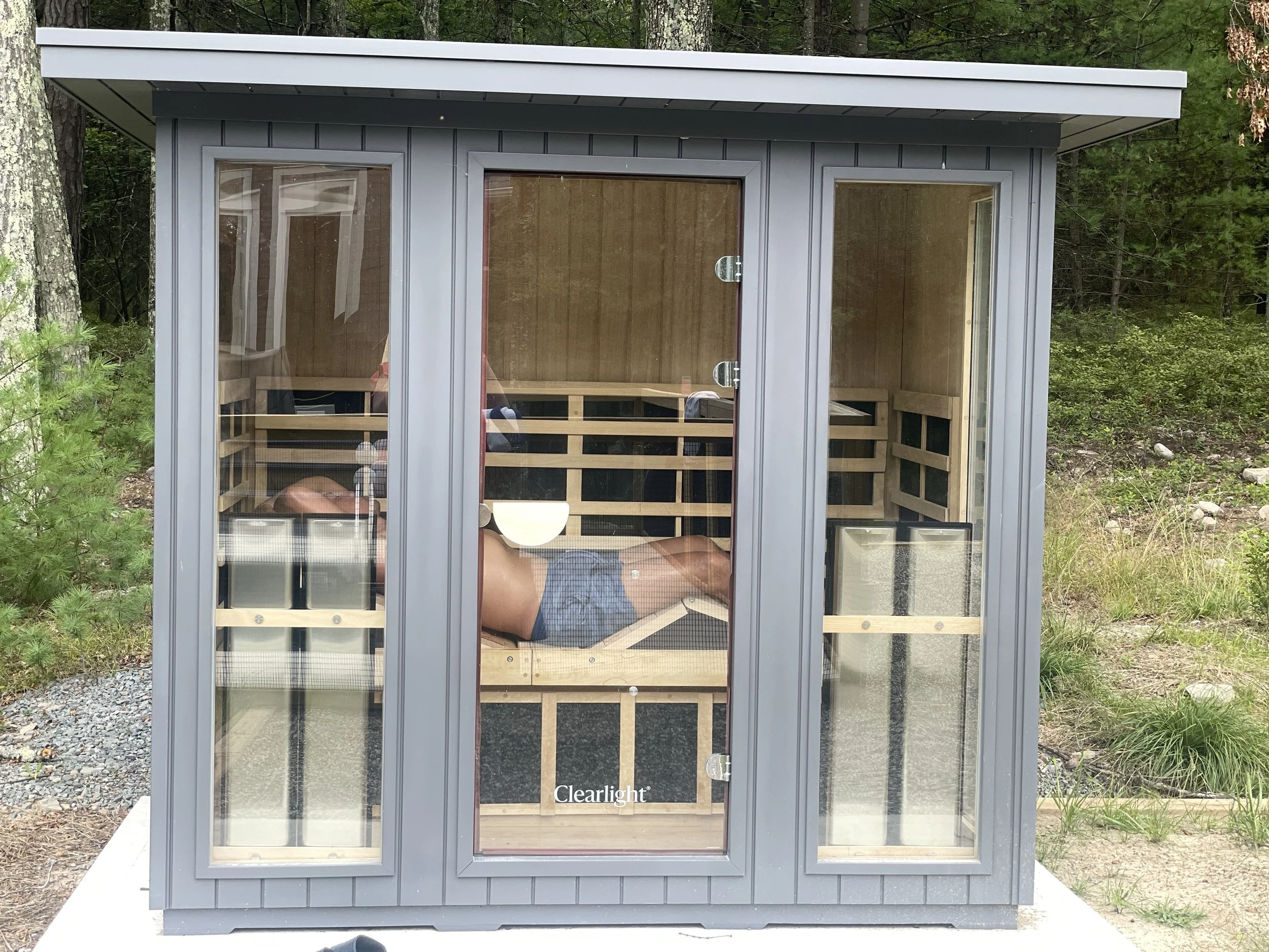 A person lying inside a wooden sauna with glass walls, situated outdoors with trees and grass around.