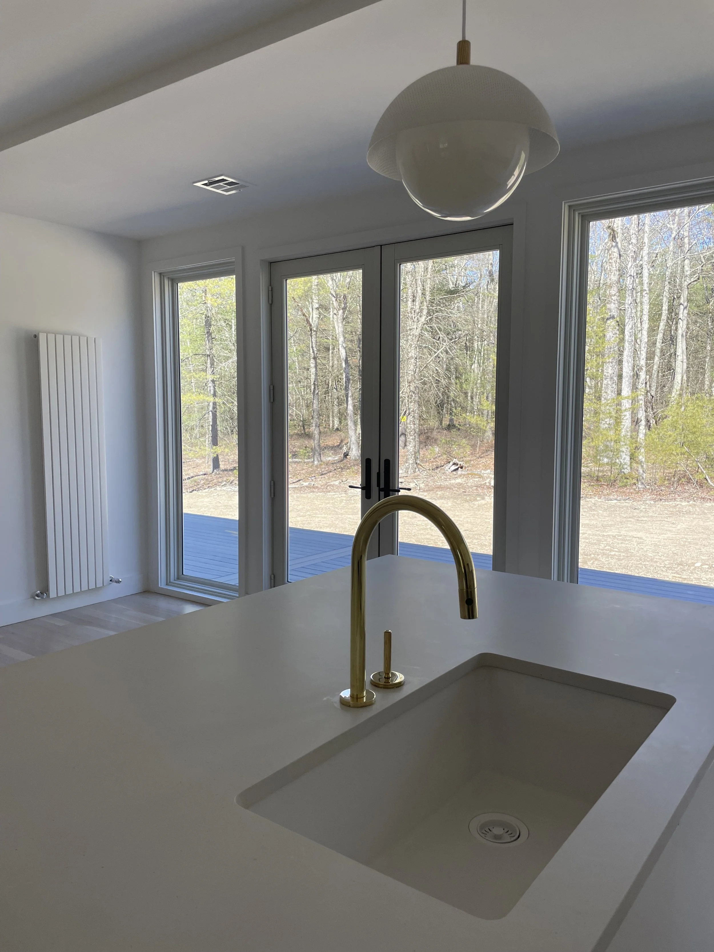 Modern kitchen with white countertops, a gold faucet, large glass sliding doors leading to an outdoor deck, and views of a wooded backyard.