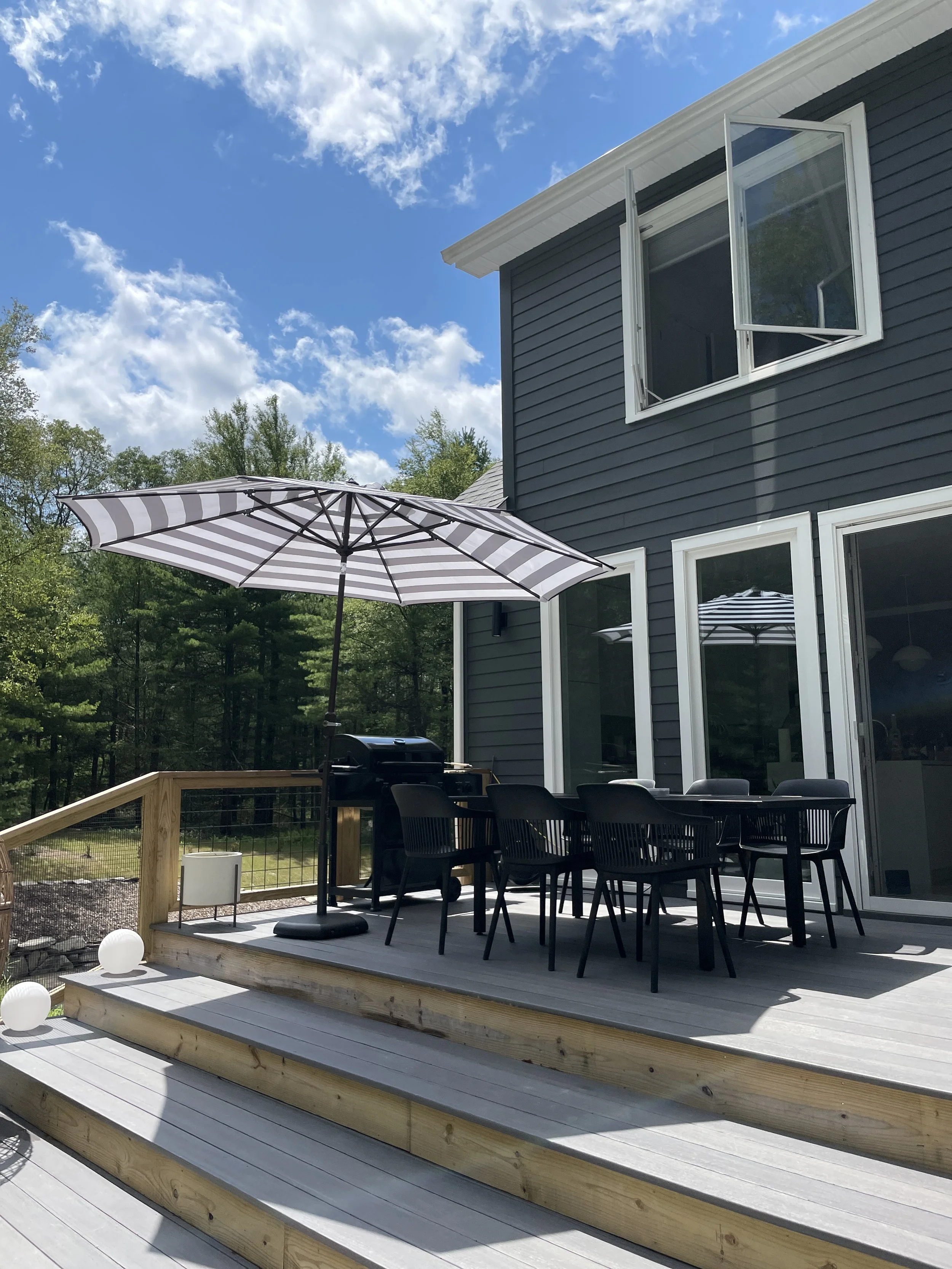 Backyard deck with outdoor dining table, chairs, barbecue grill, and striped umbrella, attached to a blue house with large glass doors, under a partly cloudy sky surrounded by trees.