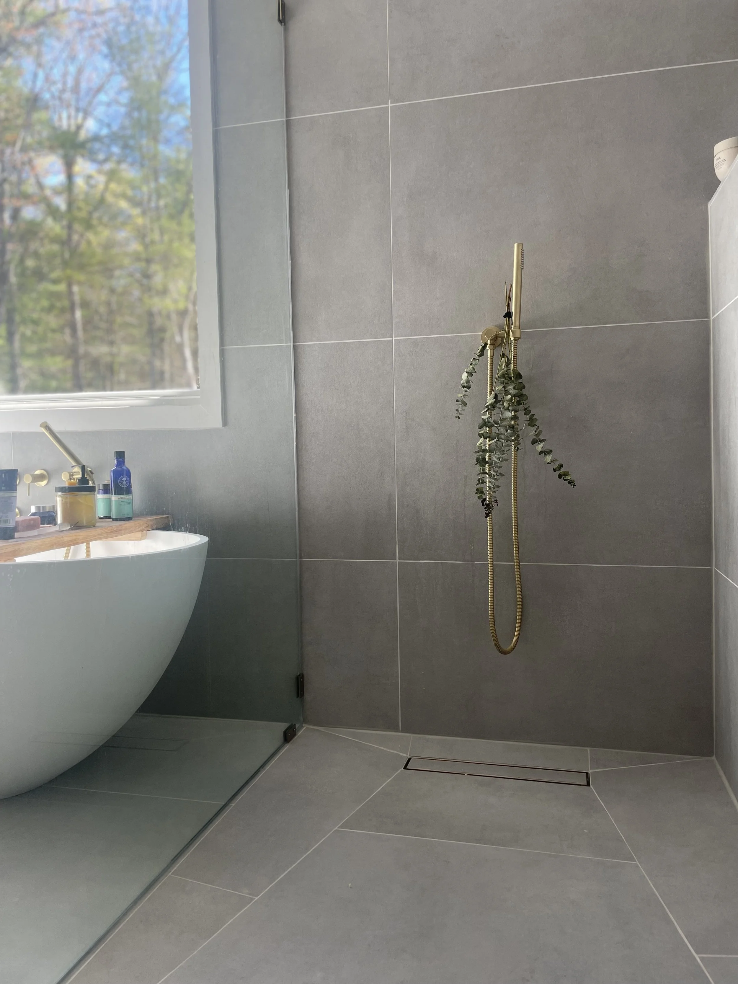 Minimalist modern bathroom with large gray tiles, a window with outdoor view, and a walk-in shower with a gold showerhead and hanging eucalyptus branches.