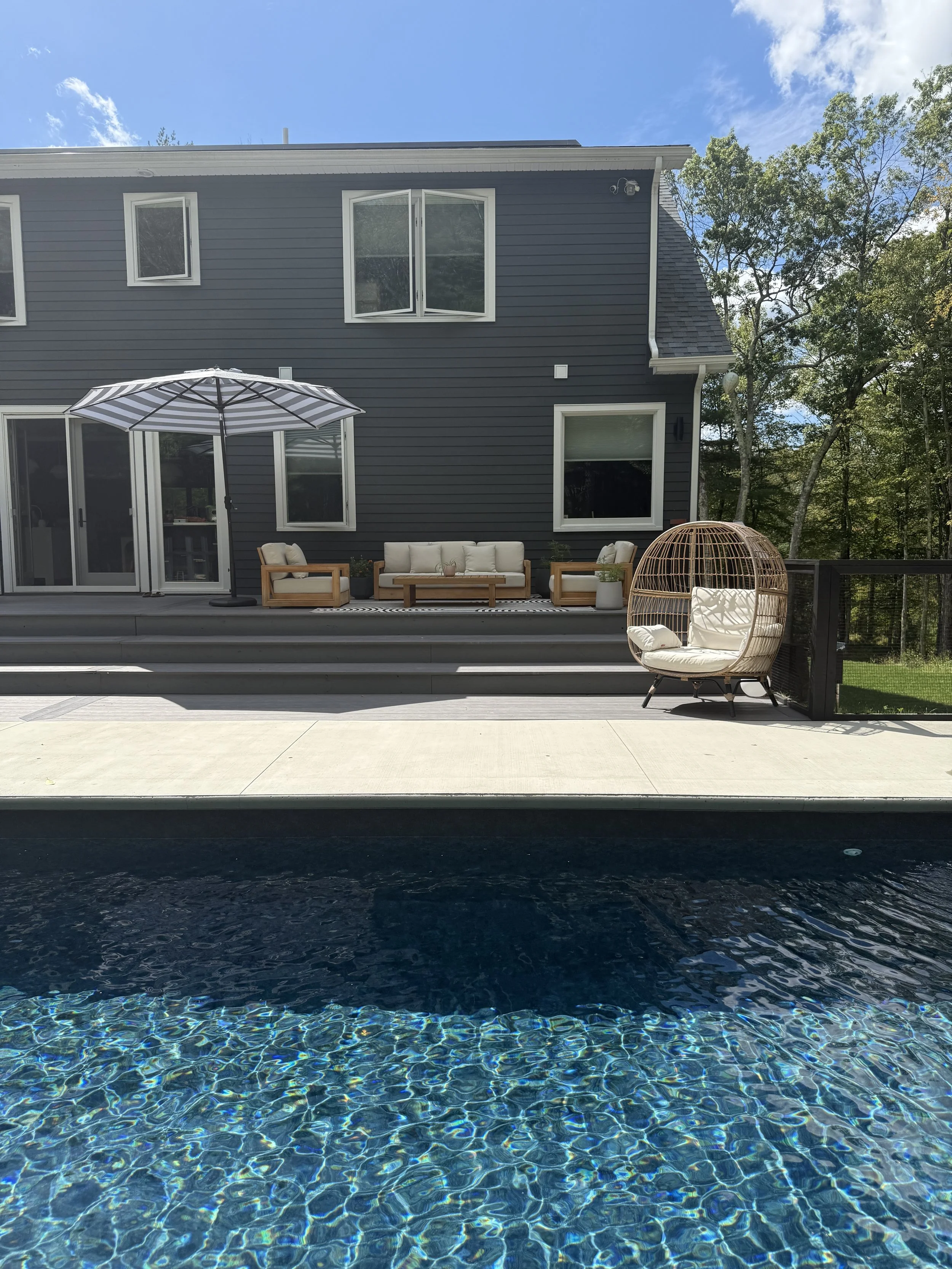 Backyard patio with a swimming pool in the foreground, a gray house with large windows and sliding glass door, outdoor furniture including a white cushioned chair with a rounded wicker cover, a seating area with a beige sofa and chairs, and a large s