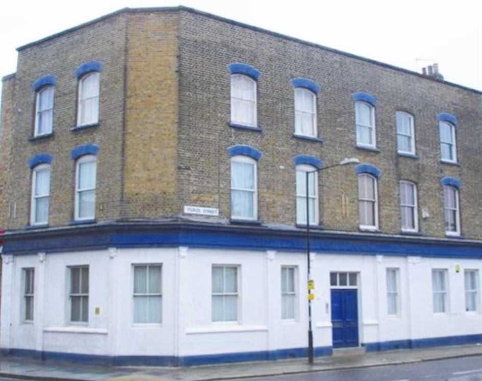 A three-story brick building on a street corner with a white lower section, blue window and door trim, and four windows on each floor with white curtains, in an urban setting.