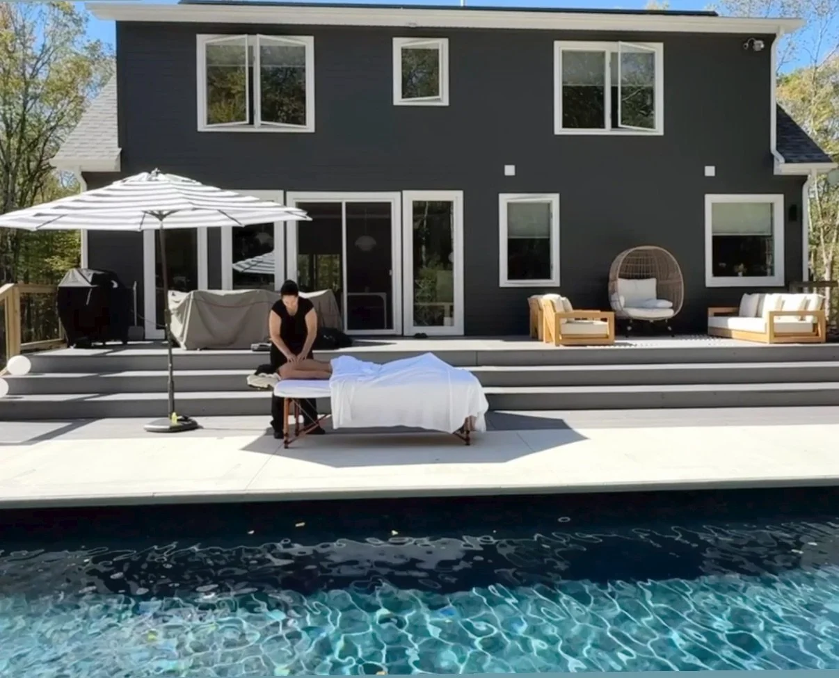 Person receiving a massage by a poolside at a modern house with outdoor furniture and a large patio umbrella.