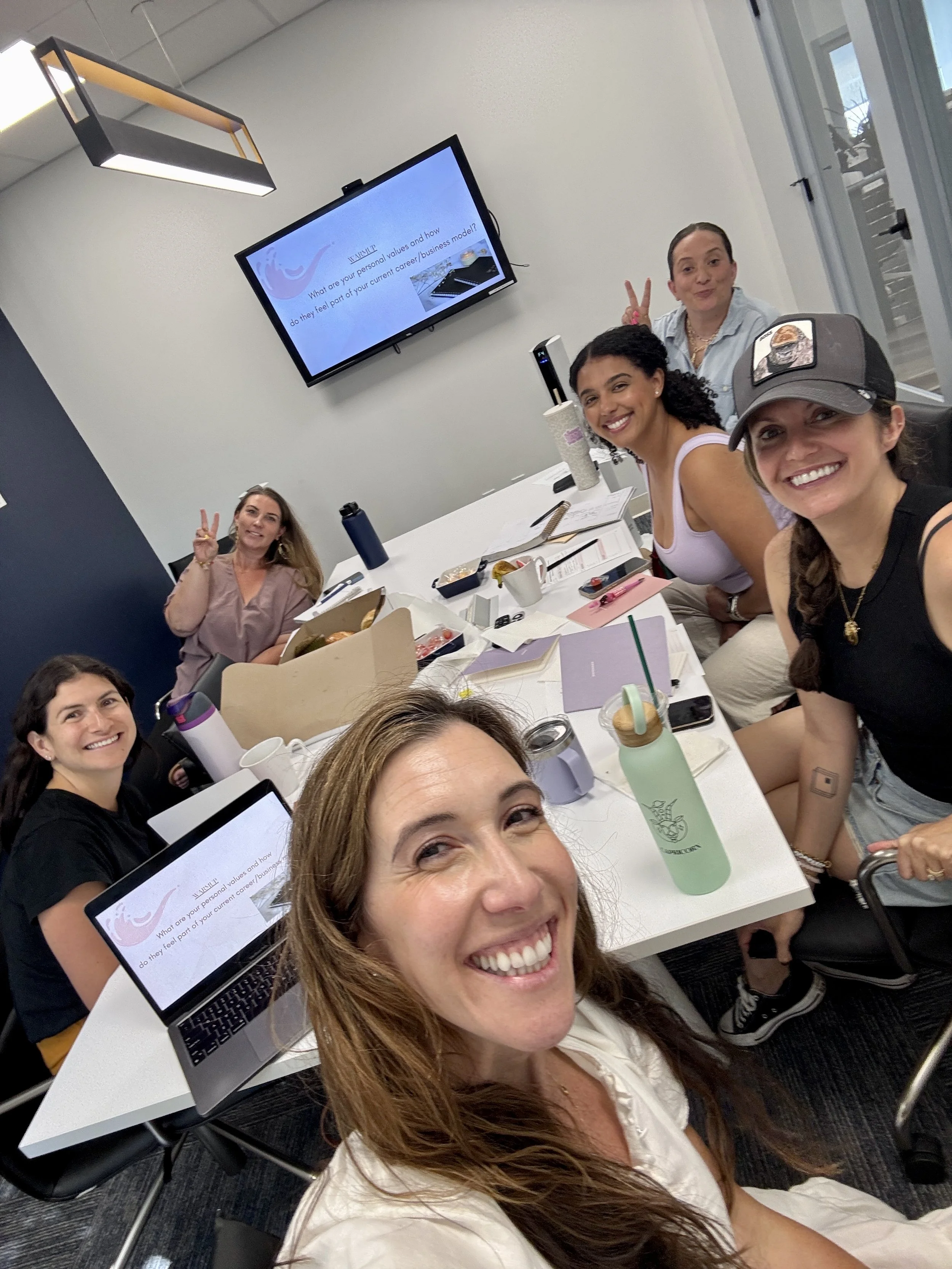 Group of smiling women sitting around a conference table in a meeting room, some making peace signs, with laptops, notebooks, drinks, and snacks on the table. A large screen displays a presentation.