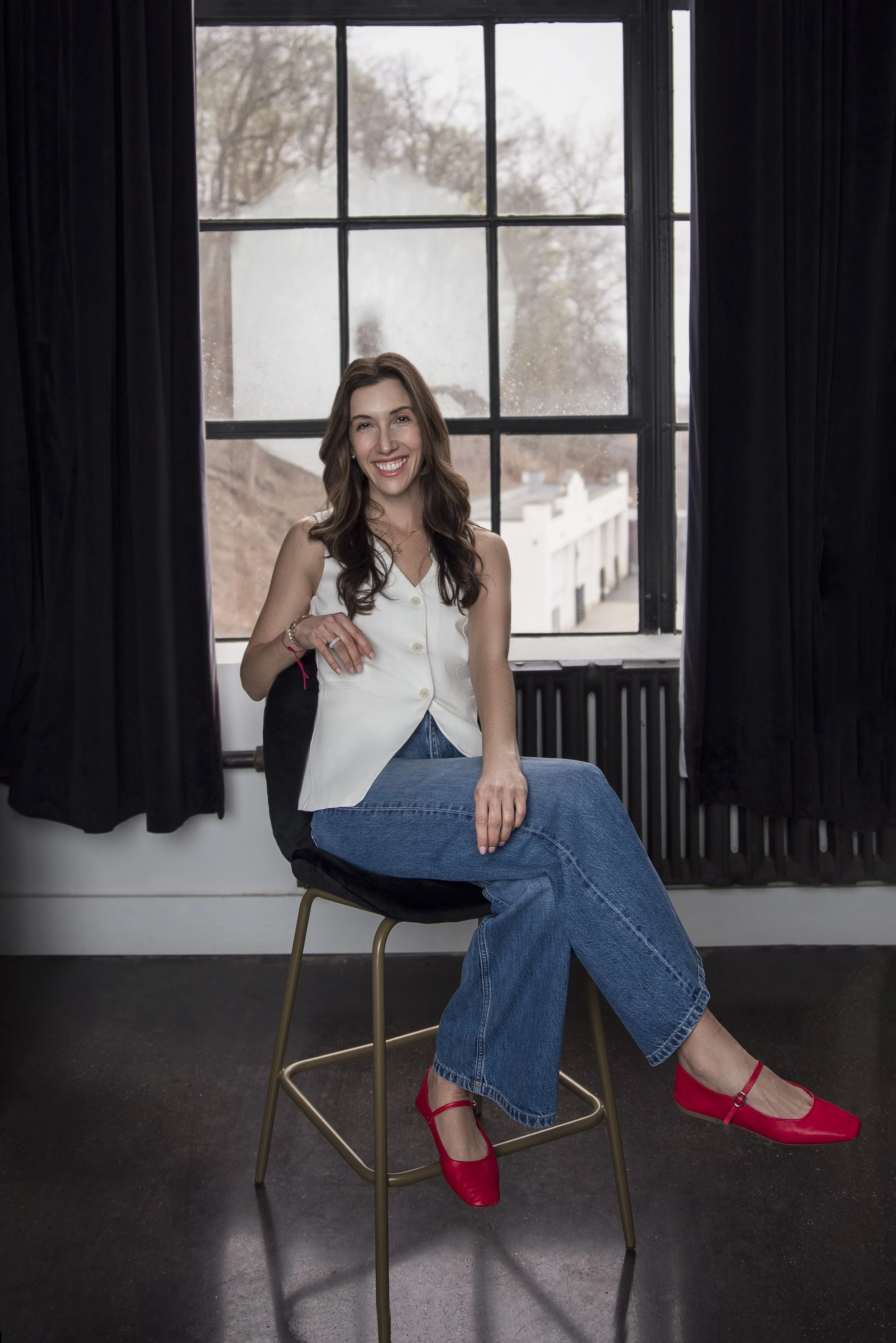 A woman with long brown hair sitting on a chair in front of a window with black curtains, wearing a white sleeveless top, blue jeans, and red shoes, smiling.