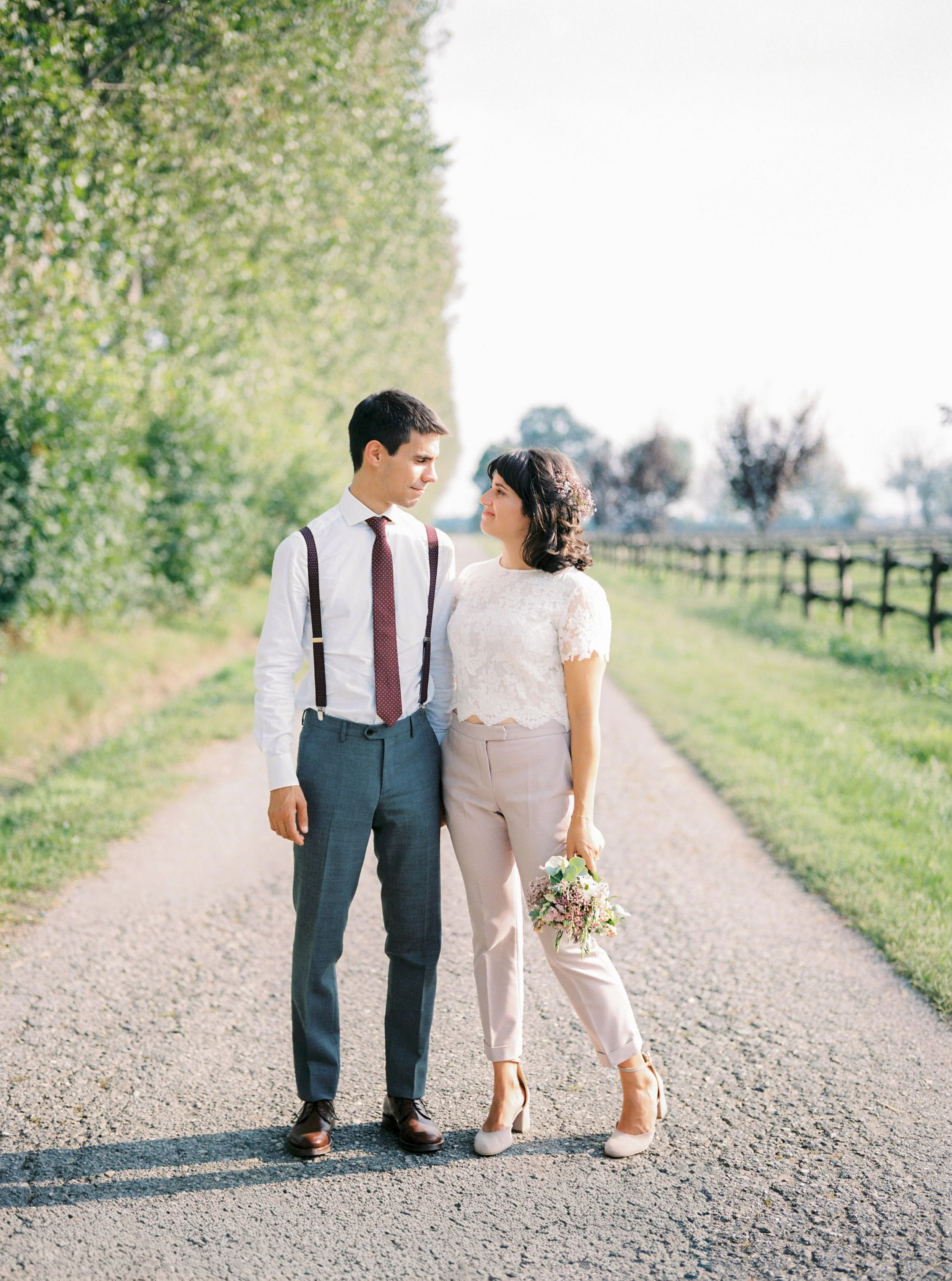 Una coppia di adulti giovani si guarda negli occhi lungo una strada di campagna, con alberi verdi ai lati e un cielo sereno.