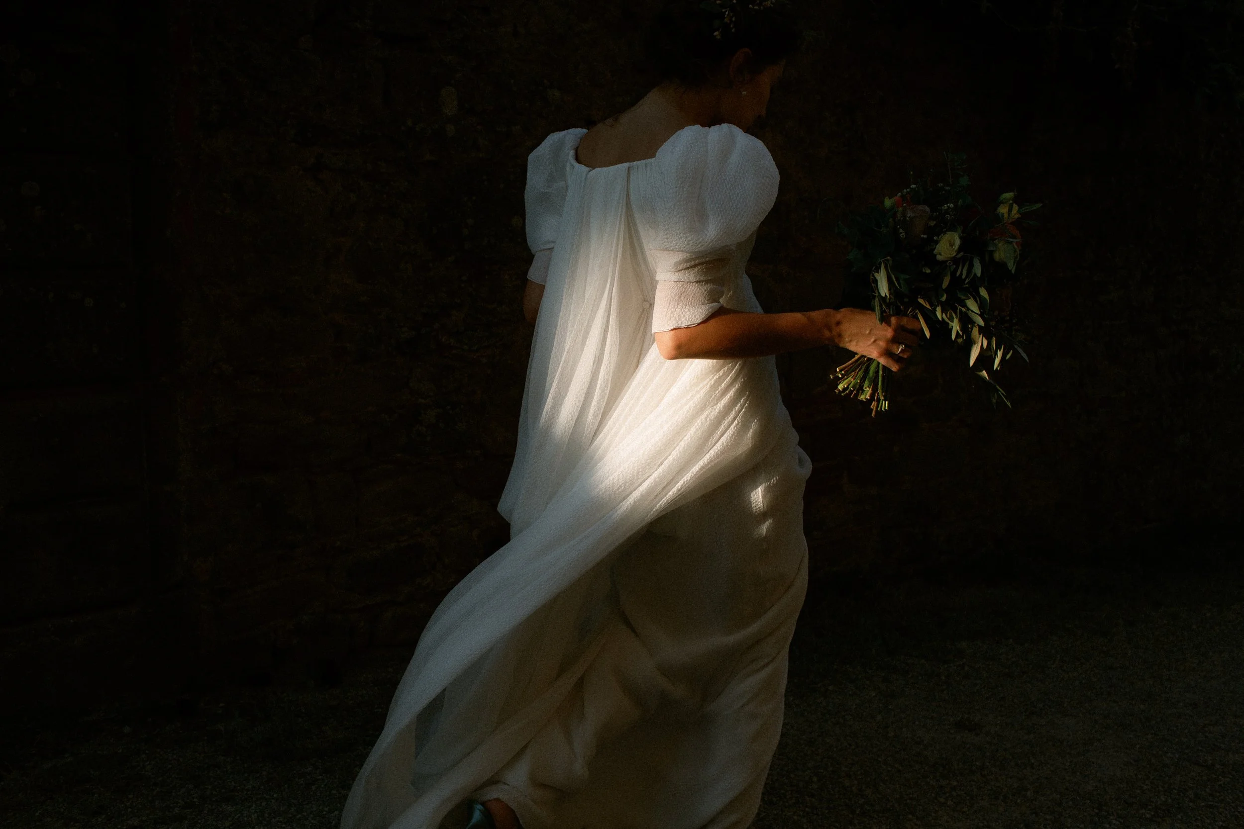 Una donna in abito bianco con maniche a sbuffo che tiene un mazzo di fiori verdi e bianchi in mano, con uno sfondo di muro di pietra.