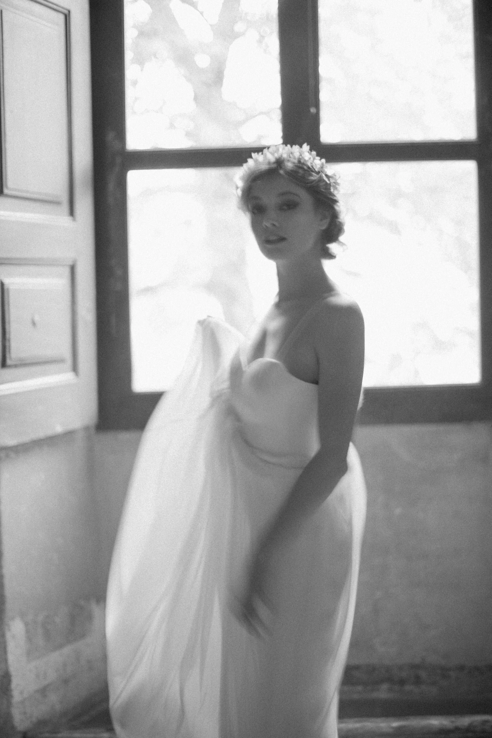 Donna in abito bianco con corona di fiori sui capelli, che si trova davanti a una finestra