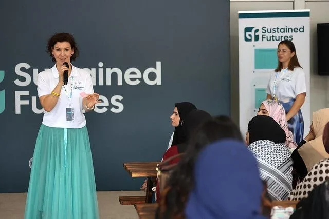 A woman in a white blouse and green skirt speaks into a microphone at a seminar or conference titled 'Sustained Futures.' Several audience members, mostly women wearing headscarves, listen attentively.
