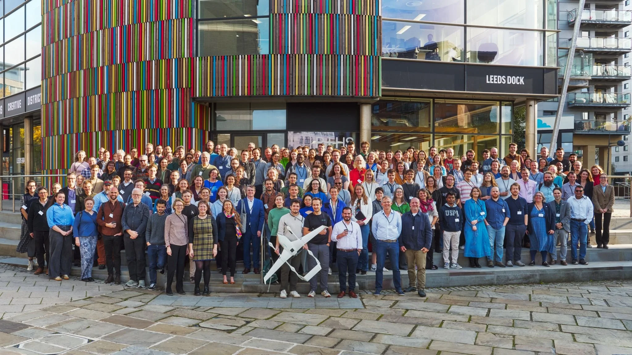 A large group of people gathered outside a modern building with colorful vertical stripes on its facade and a sign that reads 'Leeds Dock.' The group is posed for a photo, with some holding drinks and a person in the front holding a large sculpture resembling an aircraft or drone.