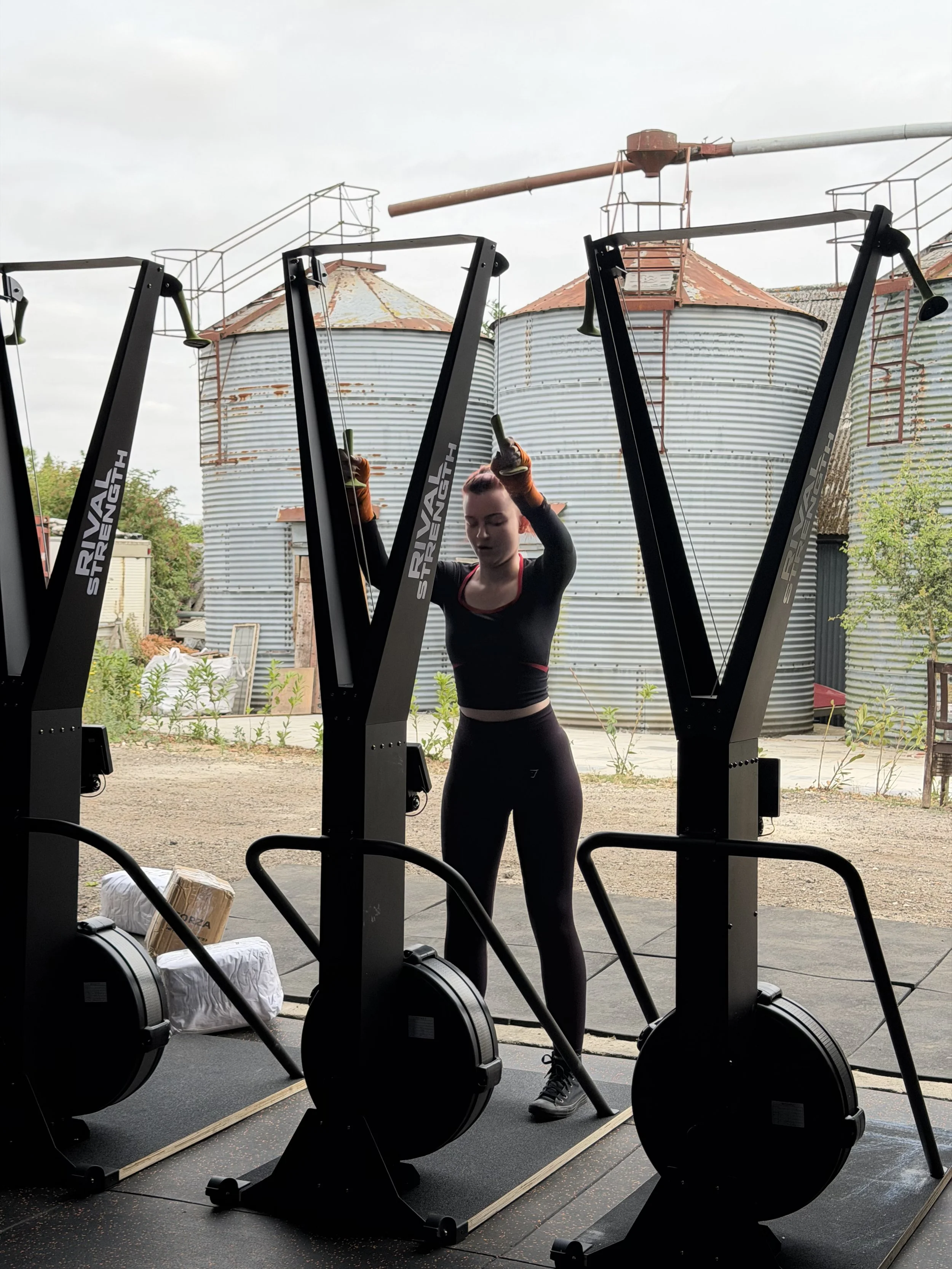 A woman standing in front of rowing machines outdoors, with farm silos and cloudy sky in the background.