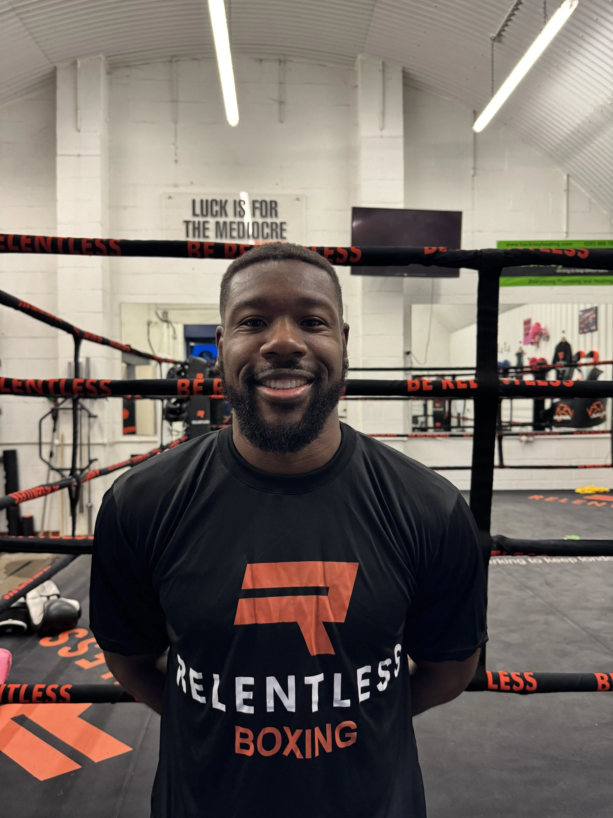 A man with tattoos on his arms smiling and sitting in a boxing gym with boxing ring ropes and a sign in the background.