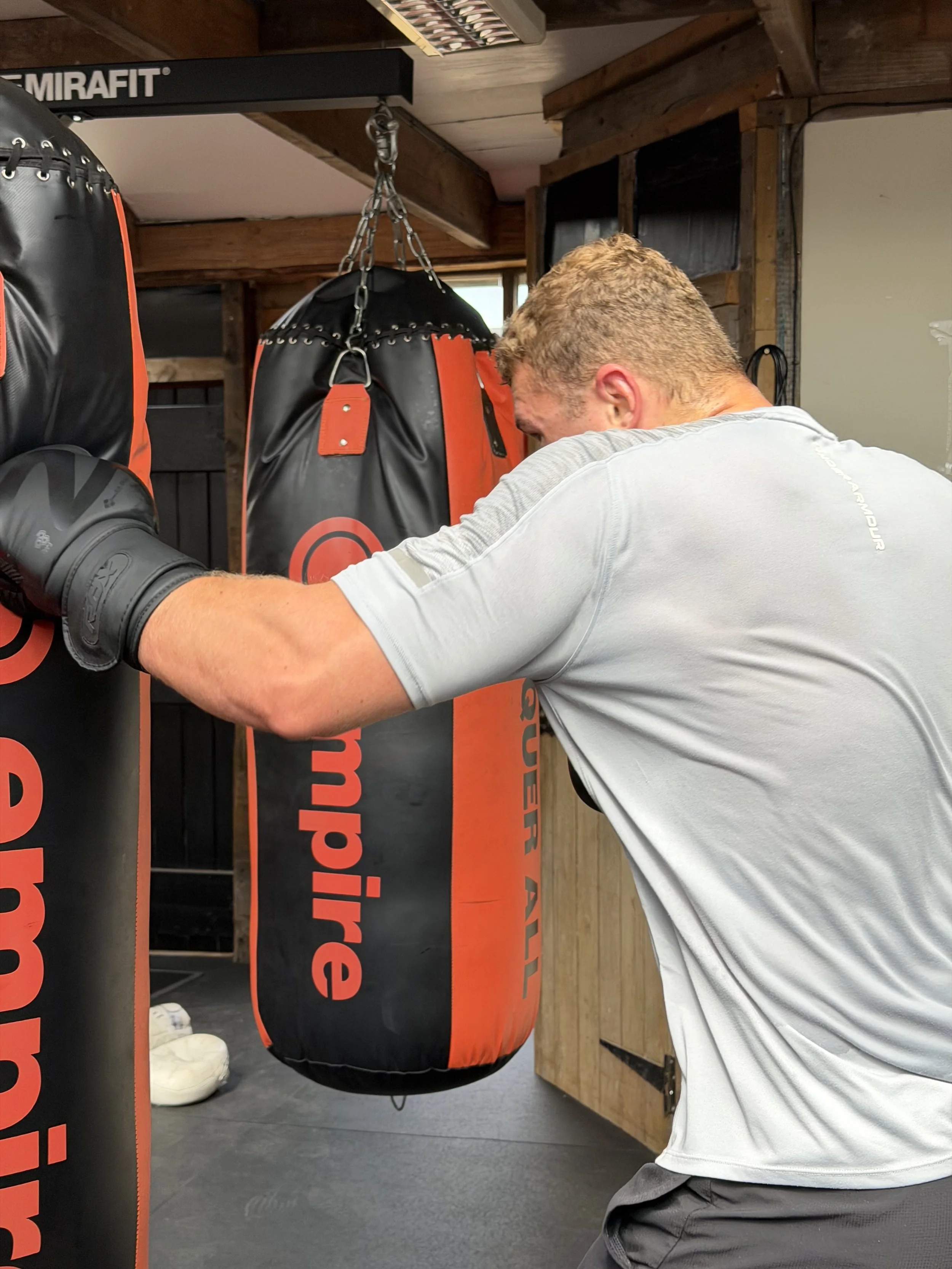 A man wearing a gray t-shirt is practicing boxing with a black and orange punching bag in a gym with wooden walls and equipment.