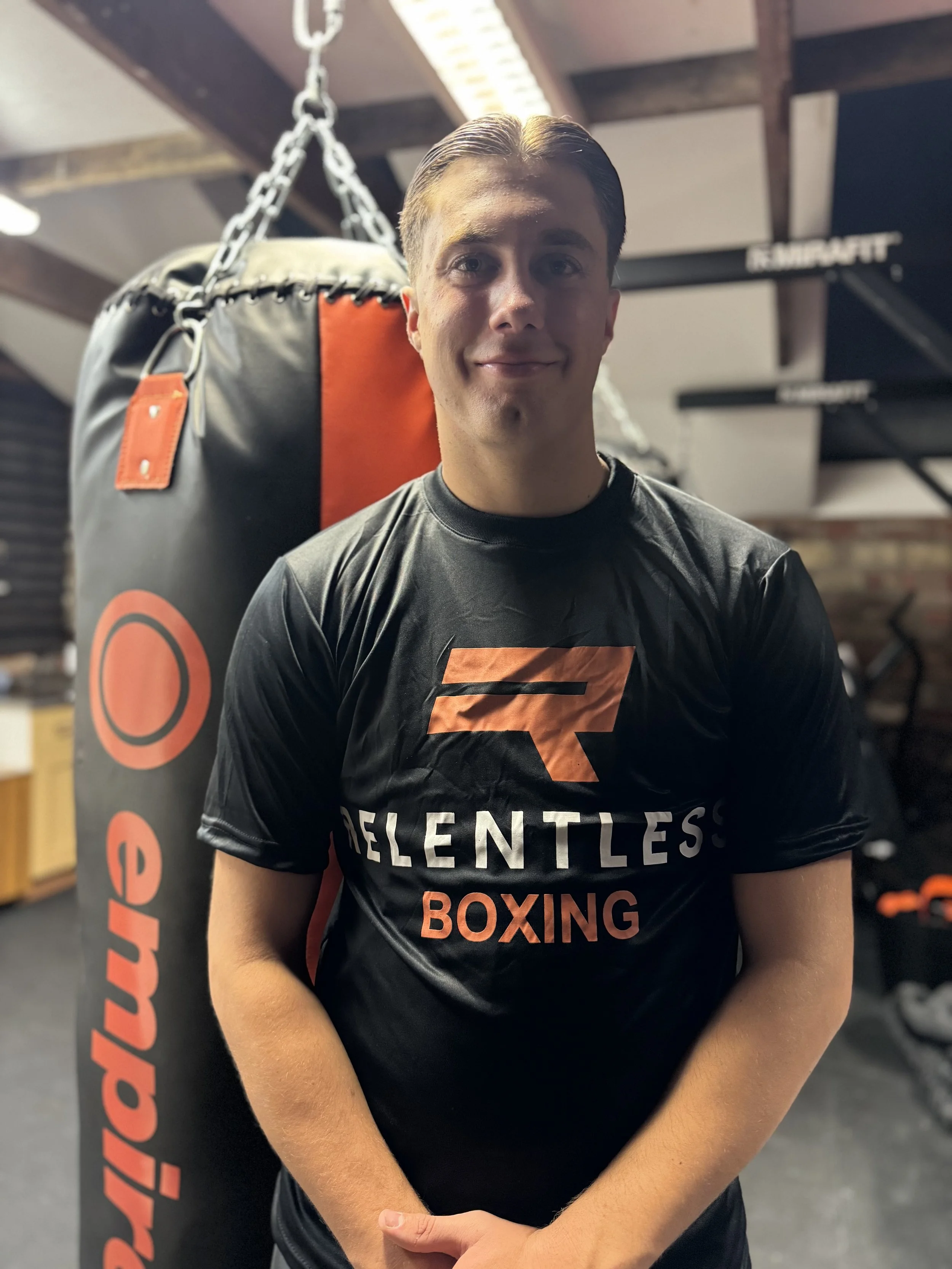A young man in a black T-shirt with the words 'Relentless Boxing' and a logo stands in a boxing gym, smiling at the camera, with a large punching bag behind him.