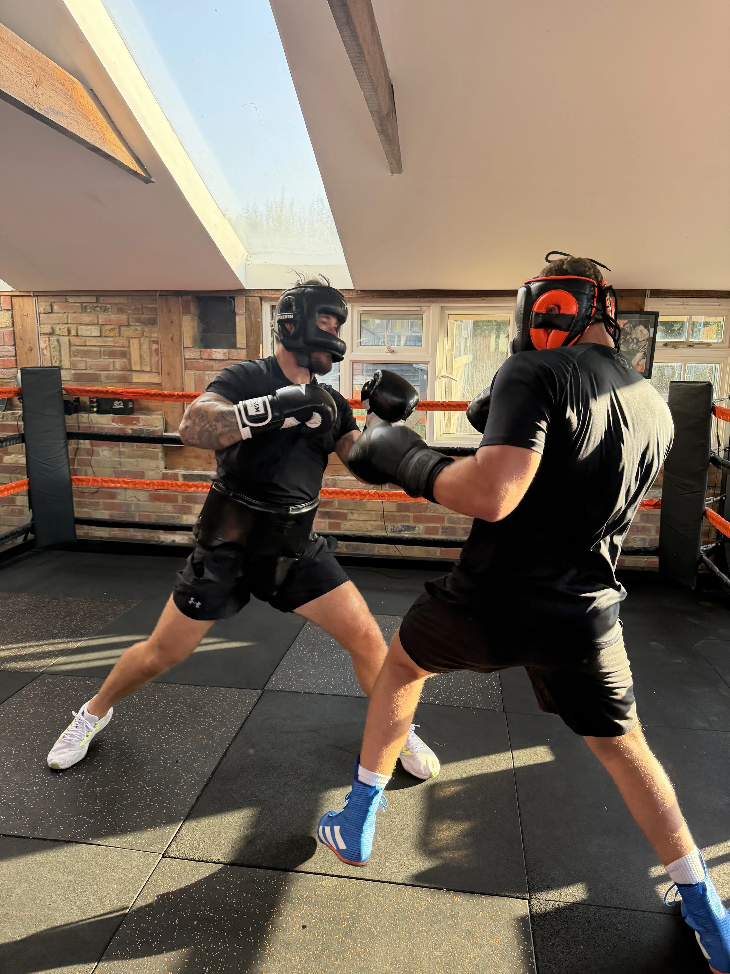 Two men engaged in boxing training inside a gym, wearing protective headgear, gloves, and athletic clothing, with boxing ring ropes visible in the background.