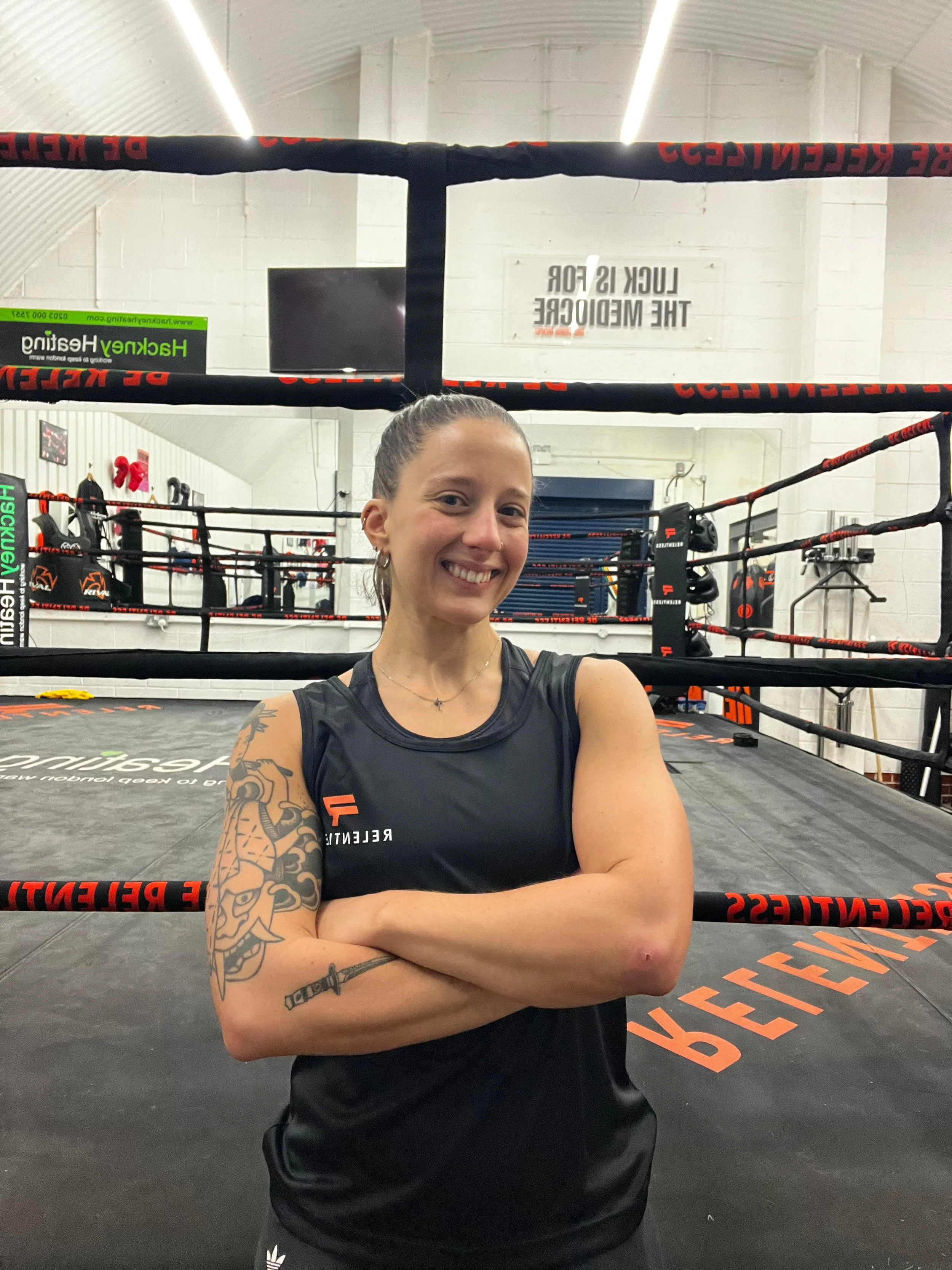 A young woman smiling in a gym, wearing a white t-shirt that says 'Be Relentless' with a pink logo, and gold jewelry.