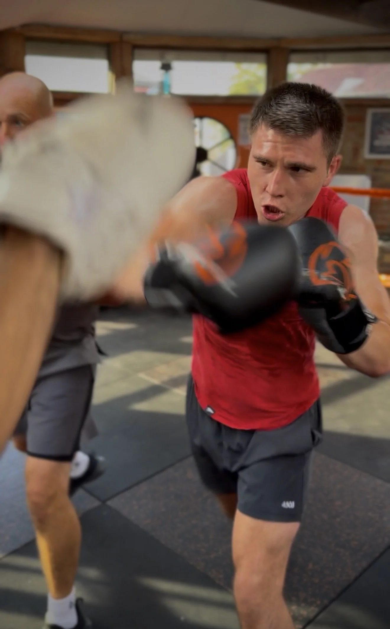 Man in red shirt and black shorts practicing boxing in a gym, with another person partially visible in the background. The man is wearing boxing gloves, and he is in an aggressive punching stance.