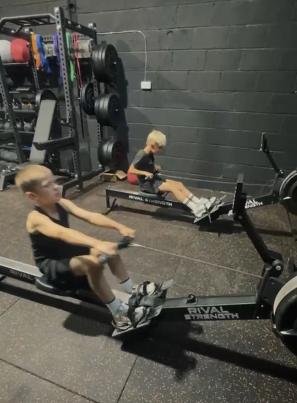 Two boys working out on rowing machines in a gym, one on an elliptical trainer and the other using a rowing machine, with gym equipment in the background.