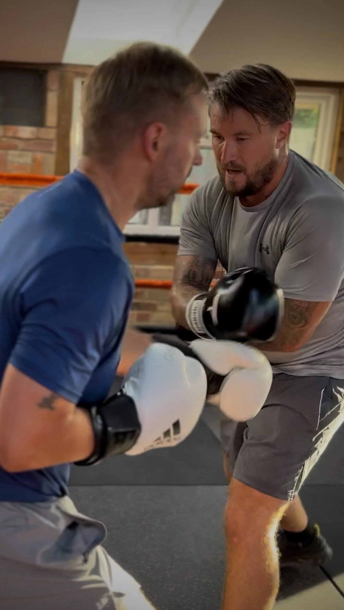 Two men training indoors, one wearing boxing gloves and the other sparring. The man with boxing gloves is dressed in a gray shirt and shorts, while the other wears a blue shirt. They are engaged in a boxing or martial arts practice, with intense focu
