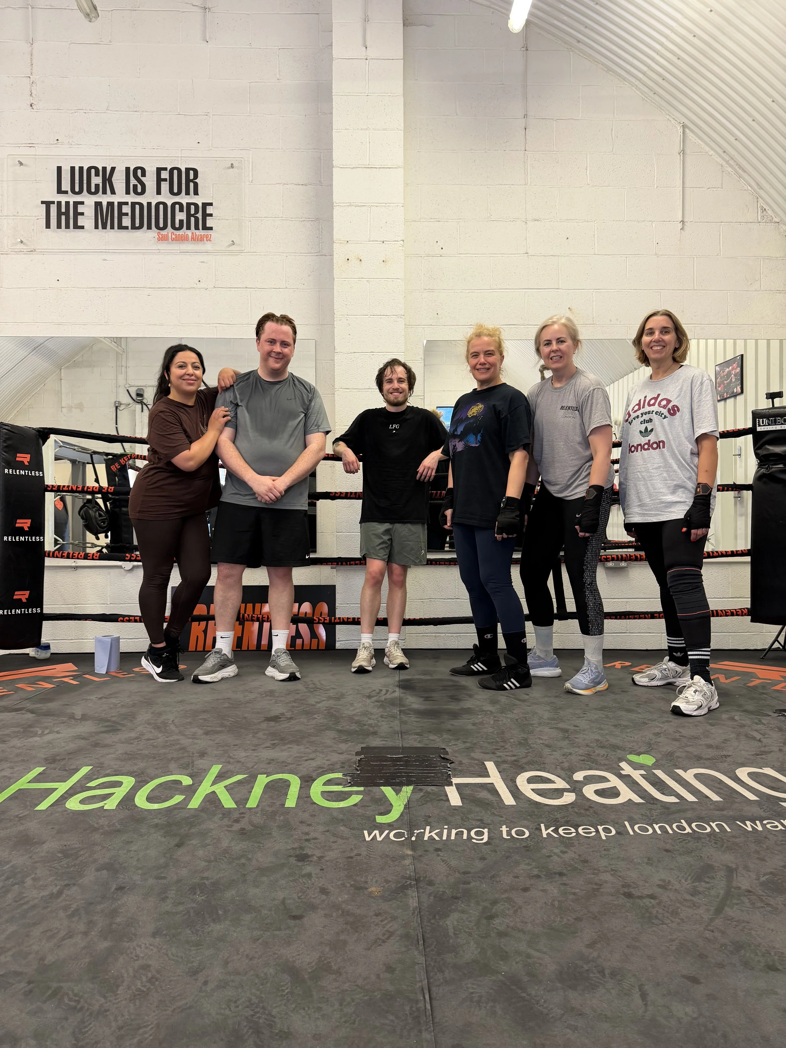 Group of six smiling people standing inside a boxing gym, posing for a photo in front of a white brick wall with a motivational quote that reads: "Luck is for the mediocre" attributed to Saul Canelo Alvarez. They are dressed in casual and workout clothing, and some are wearing boxing gloves.