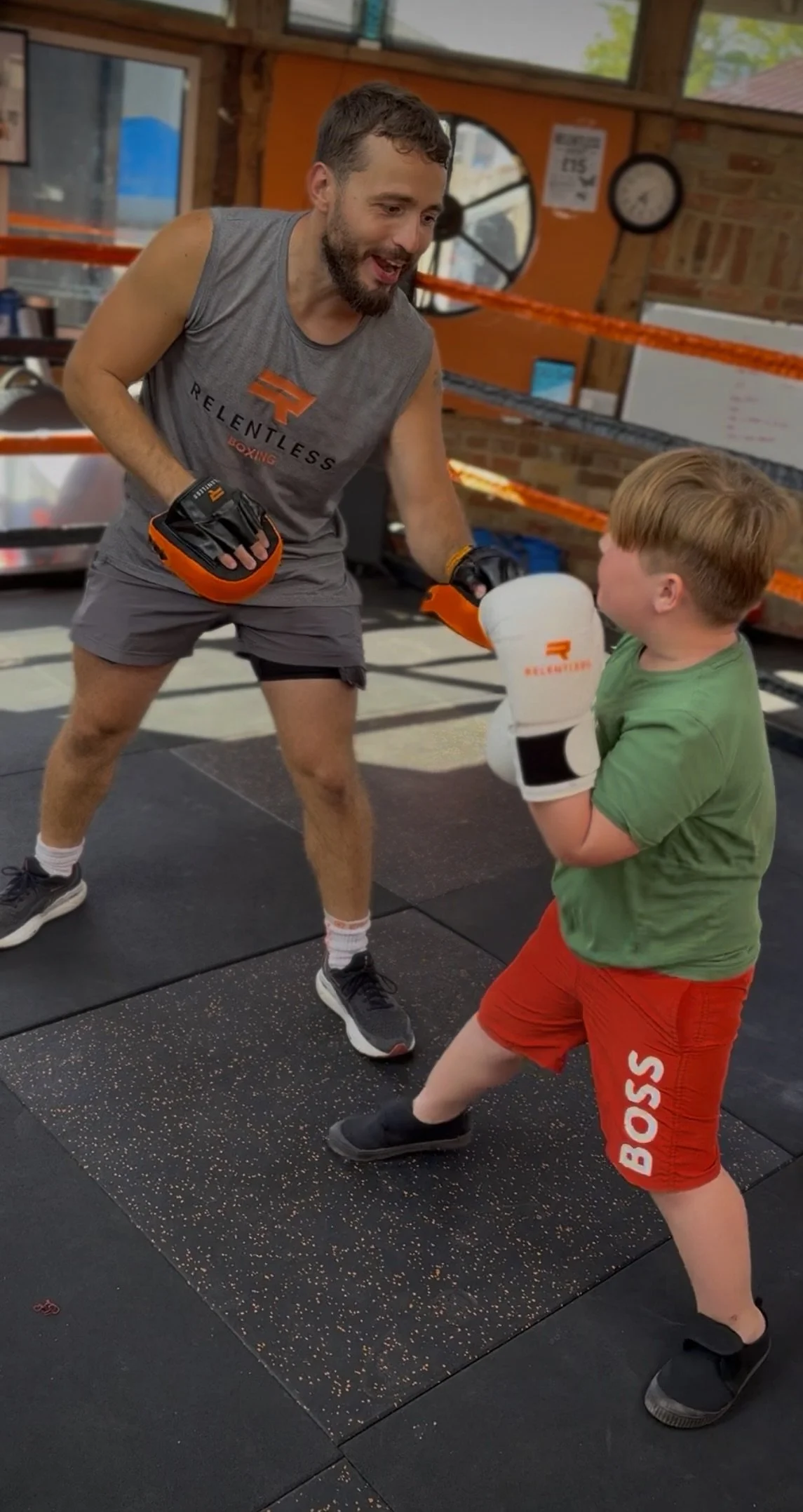 A man and a young boy sparring in a boxing gym. The man is wearing a sleeveless athletic shirt and shorts, and the boy is wearing a green T-shirt and red shorts. Both are wearing boxing gloves. The boy is throwing a punch while the man is smiling and