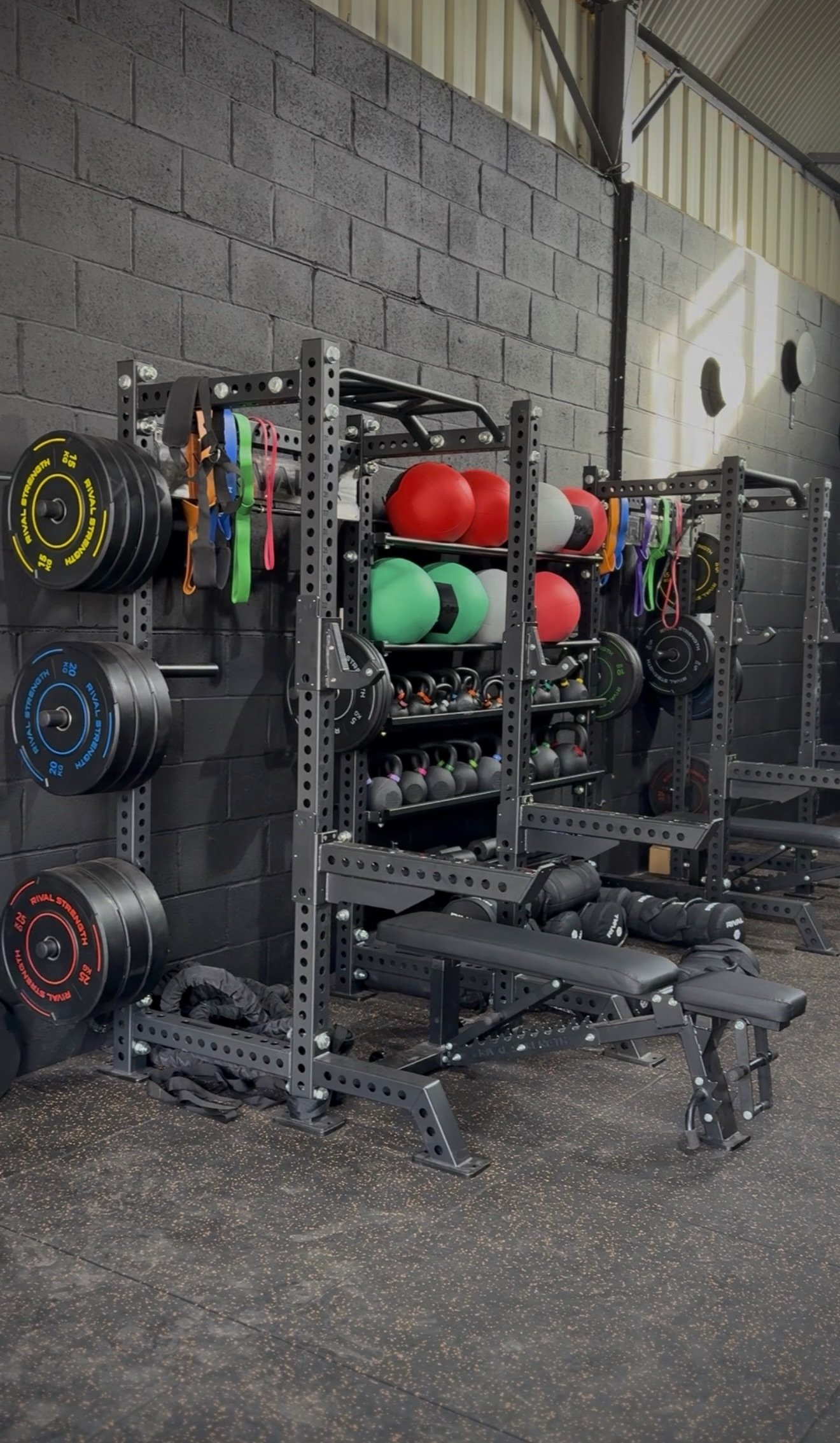 A gym rack with weight plates, medicine balls, resistance bands, kettlebells, and a workout bench against a dark brick wall.