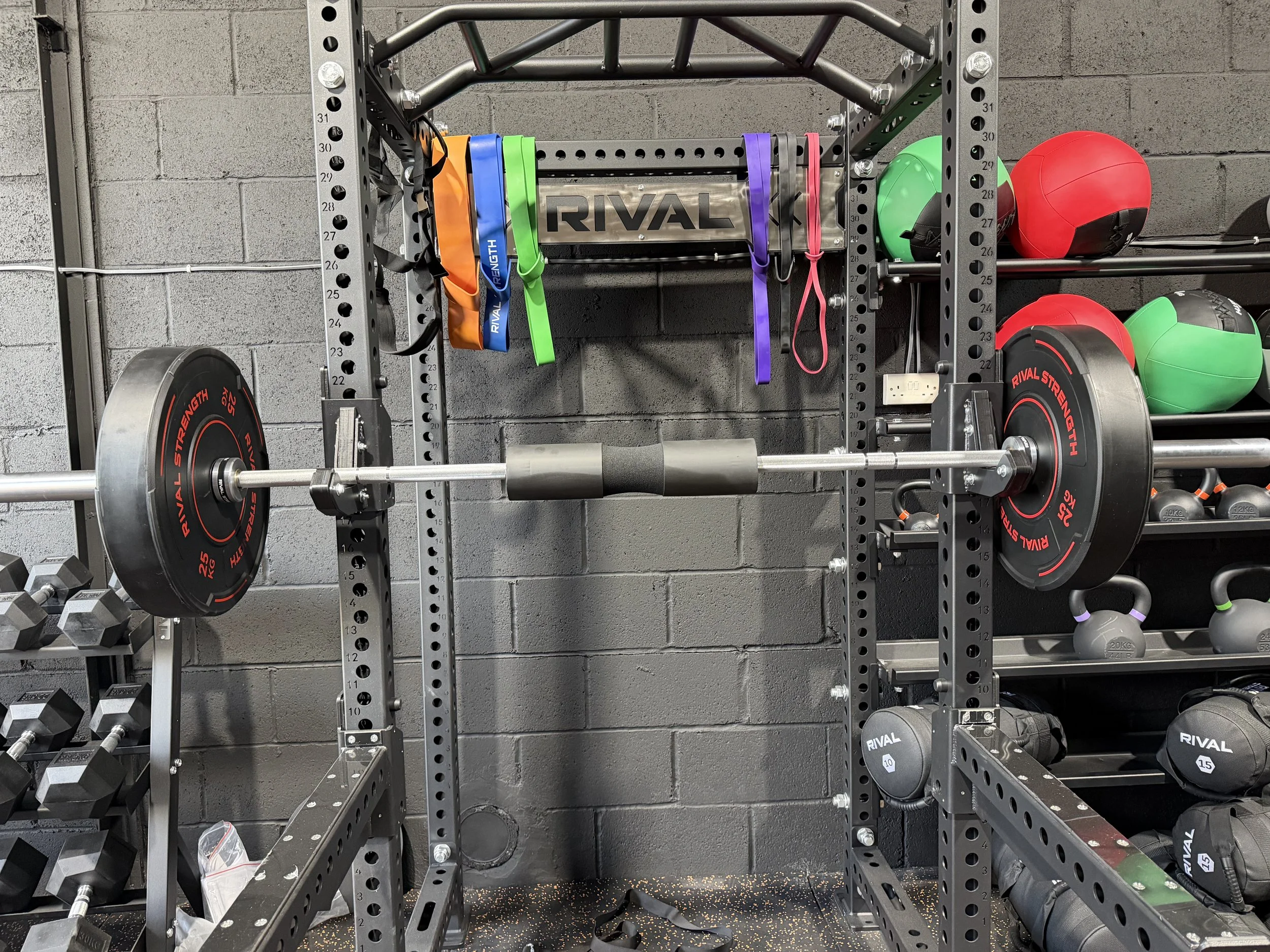 A metal workout rack holding weight plates, resistance bands, medicine balls, and kettlebells against a gray brick wall.