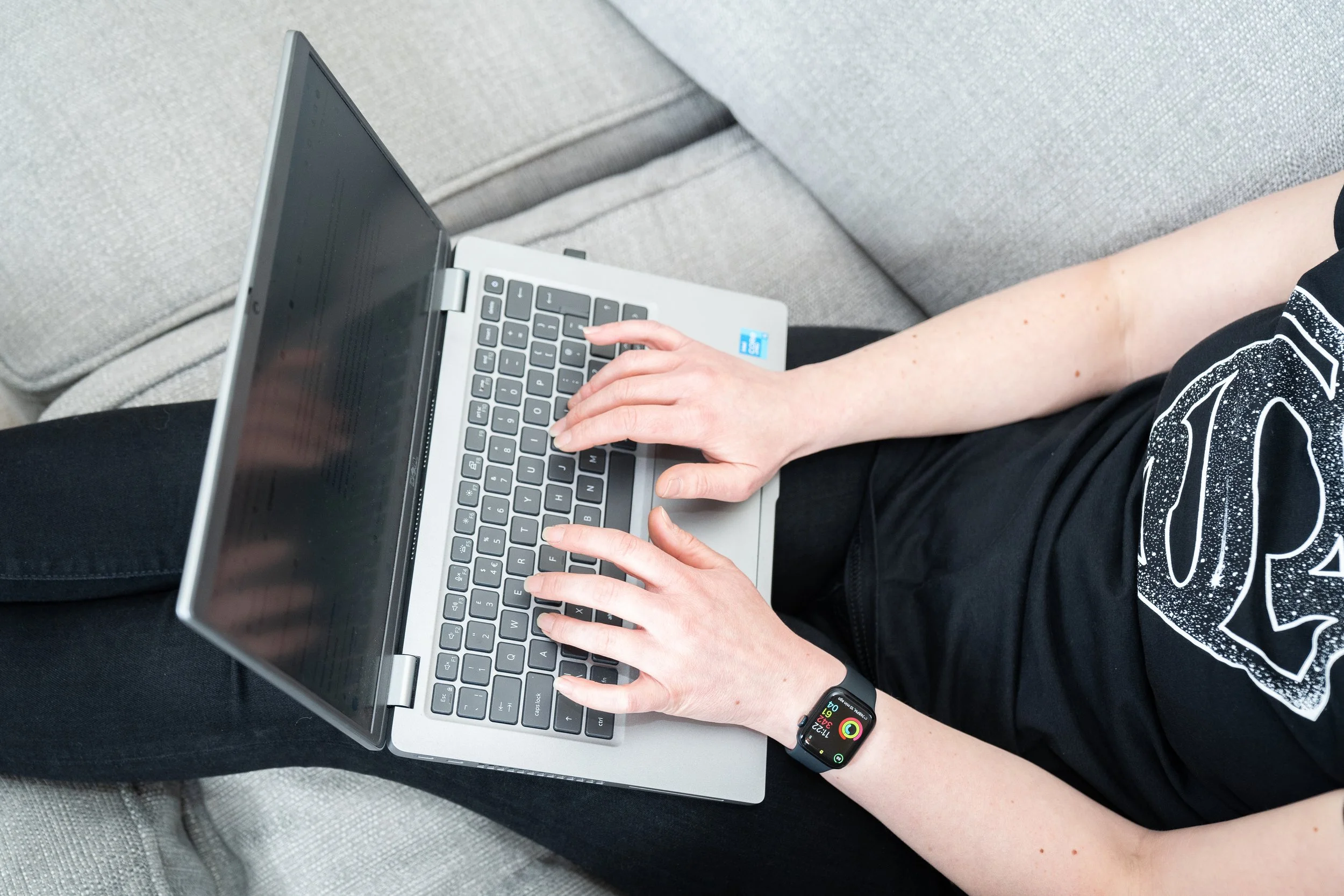 Person sitting on a couch, using a silver laptop with a black keyboard, wearing a black smartwatch, visible on their left wrist for Expert Bid Writing Service and Bid Support