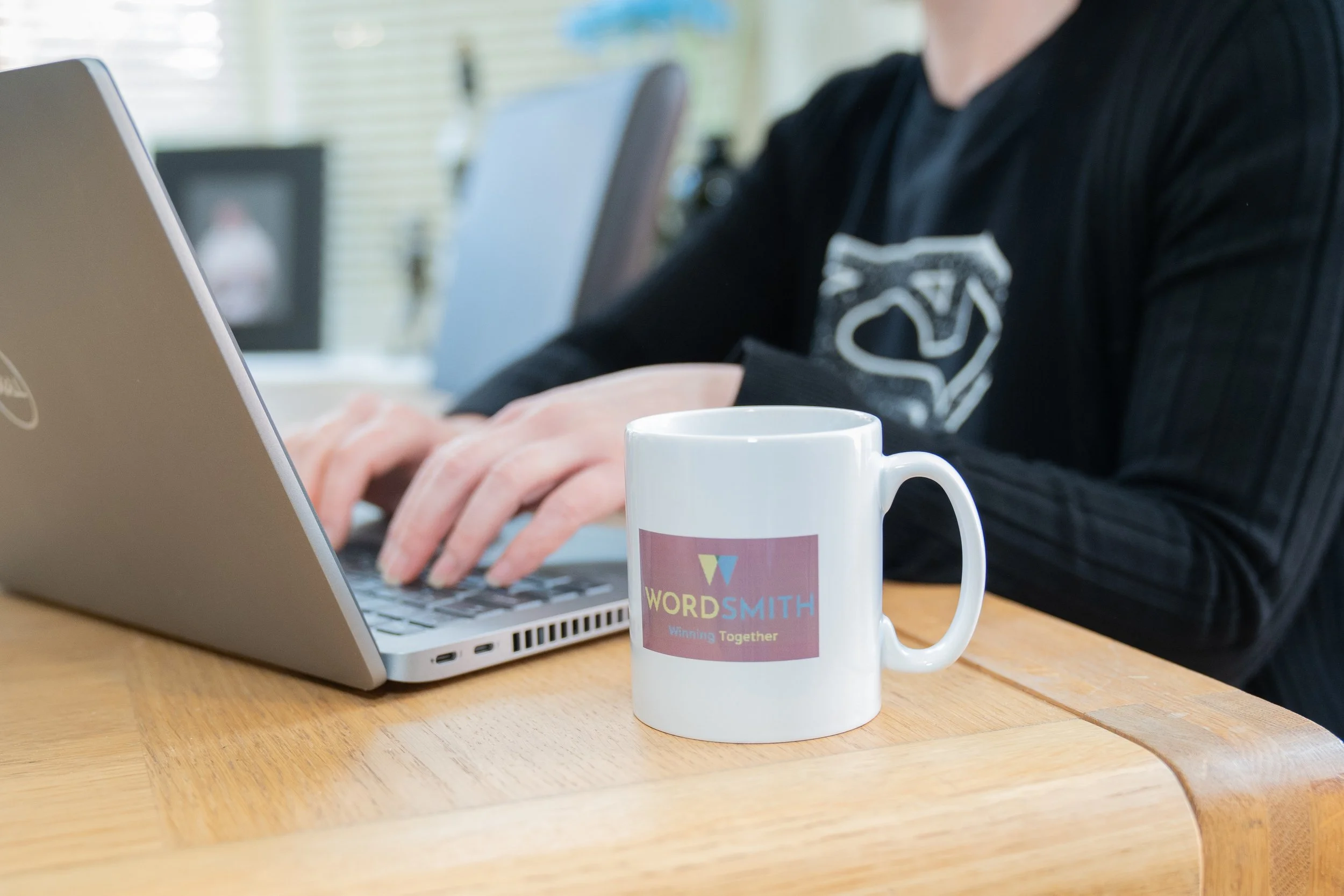 A person typing on a silver laptop on a wooden table, with a white coffee mug labeled 'WORDSMITH' in the foreground for the page Expert Bid Writing Service and Bid Support.