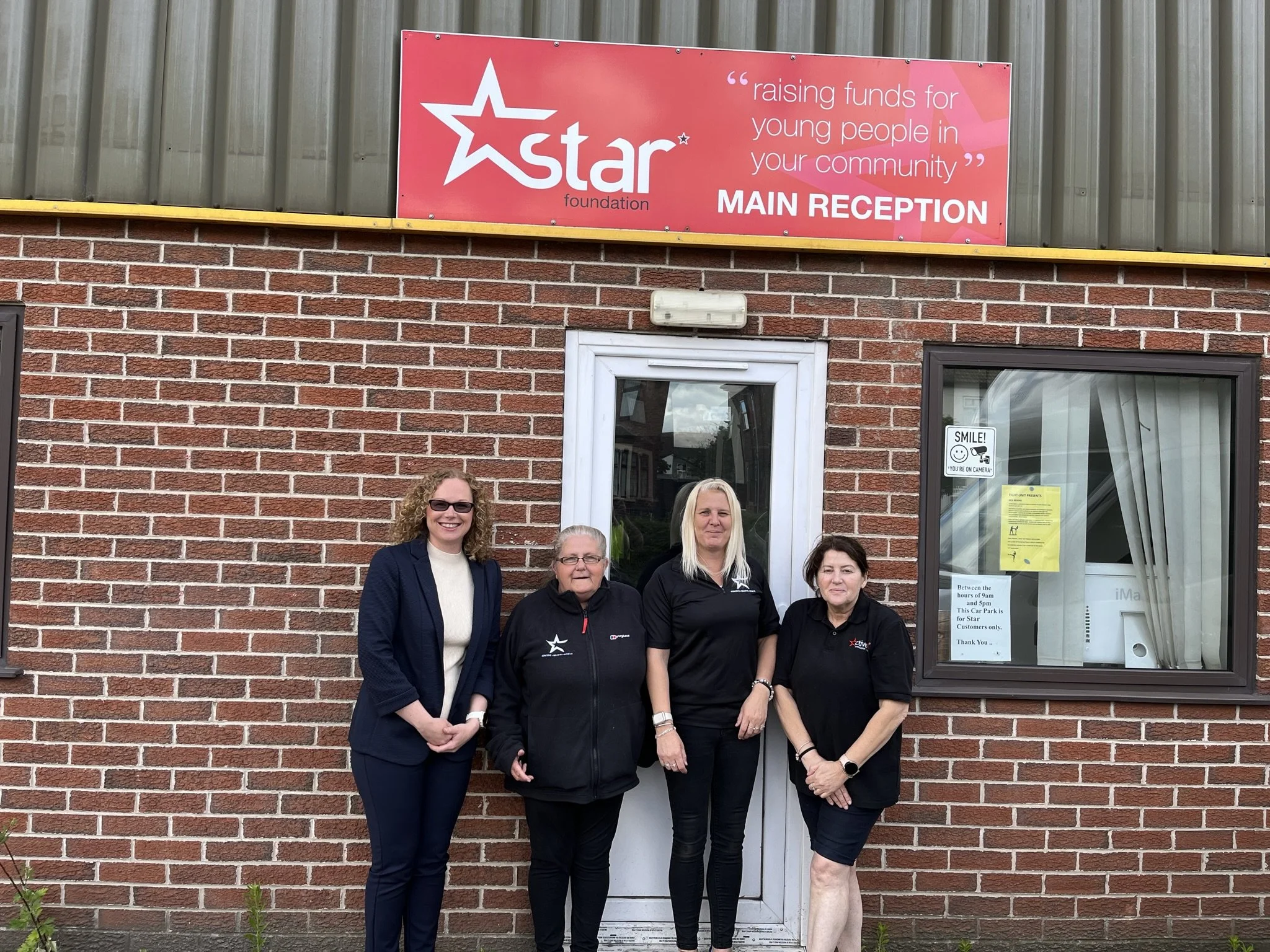 Four women standing in front of a brick building with a large red sign that reads 'Star Foundation' and 'raising funds for young people in your community.'