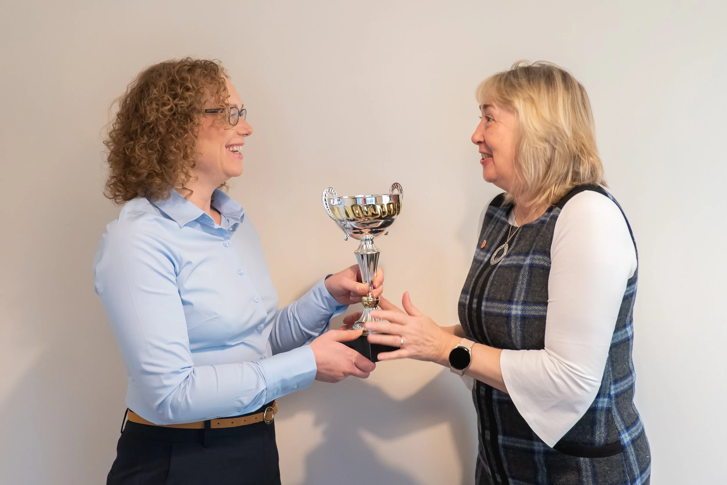 Two women smiling and exchanging a trophy indoors against a plain wall for the page Expert Bid Writing Service and Bid Support
